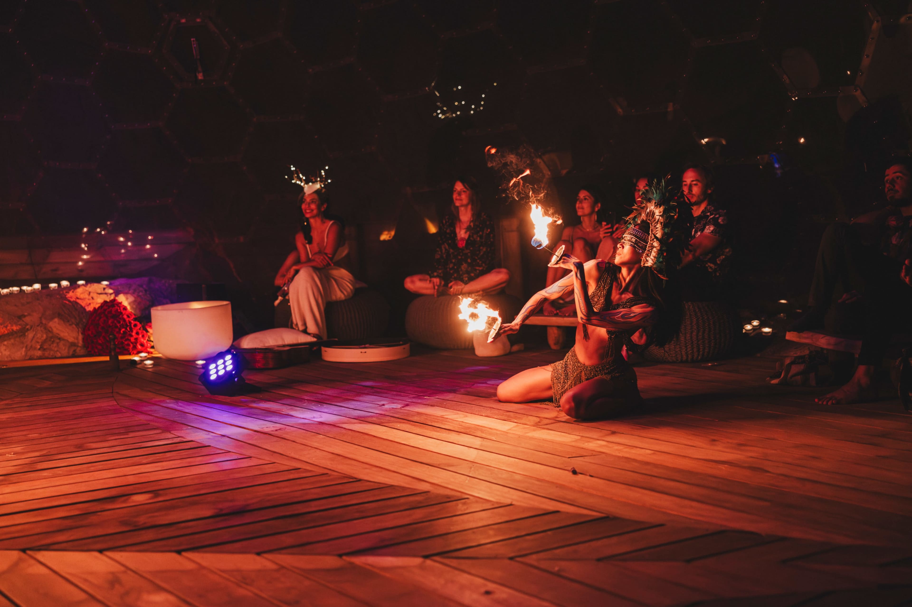 A performer dances with fire in a geodesic dome, while an audience sits on cushions, watching the performance.