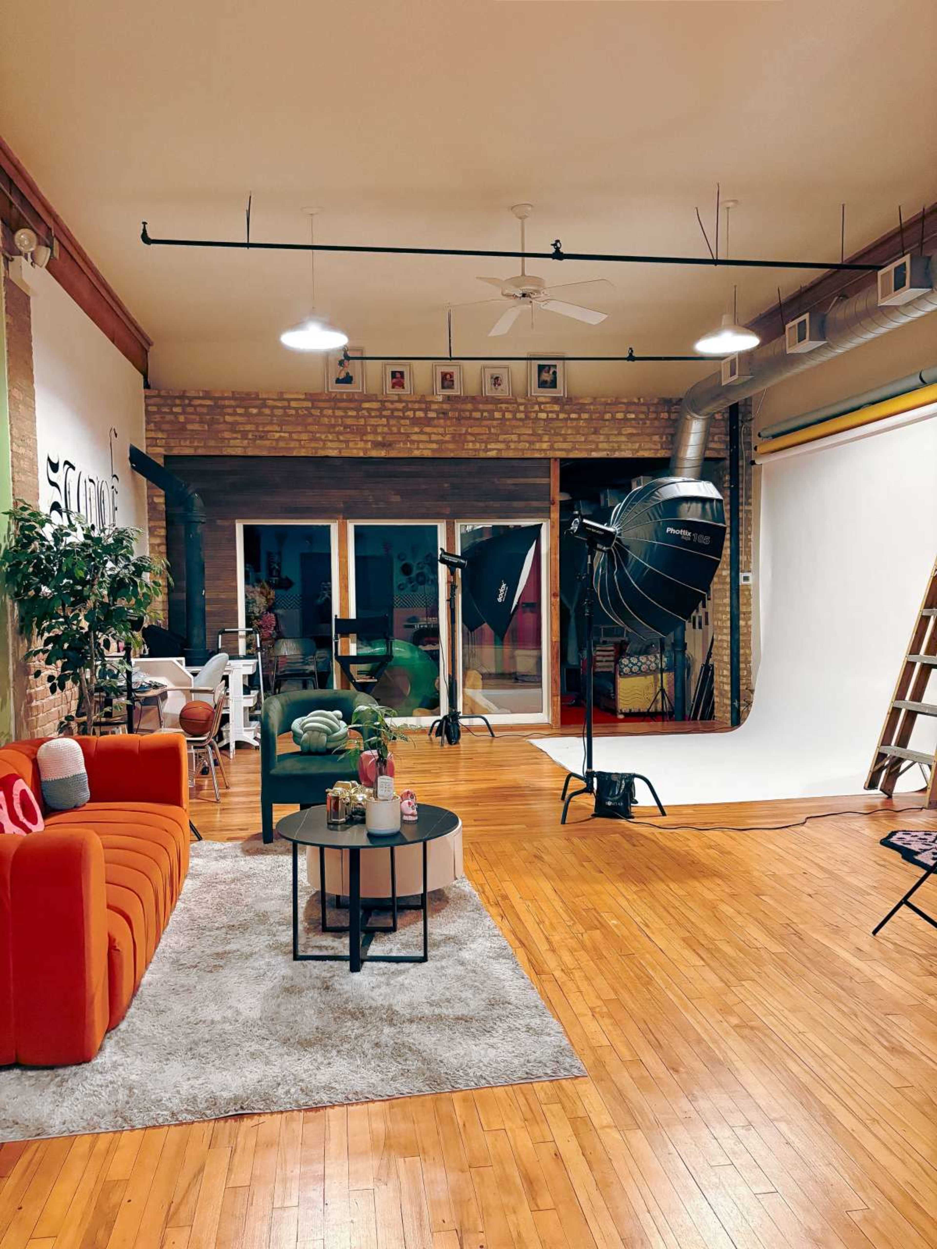 The image shows a photography studio with a large backdrop, lighting equipment, a seating area with an orange couch, and wooden flooring.