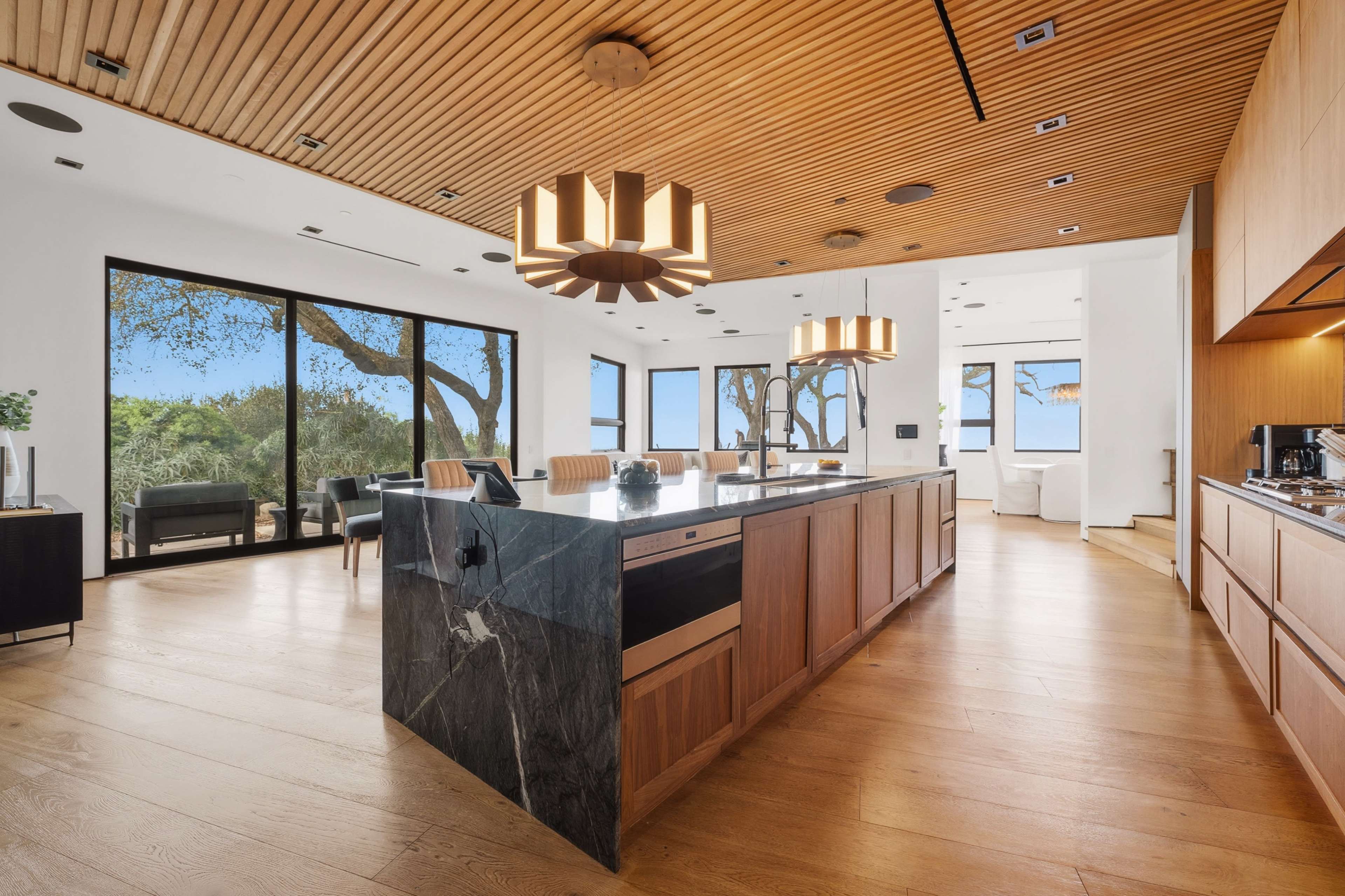 The image showcases a modern kitchen and dining area featuring a large island with a marble countertop, wooden cabinetry, and large windows that overlook a scenic view.