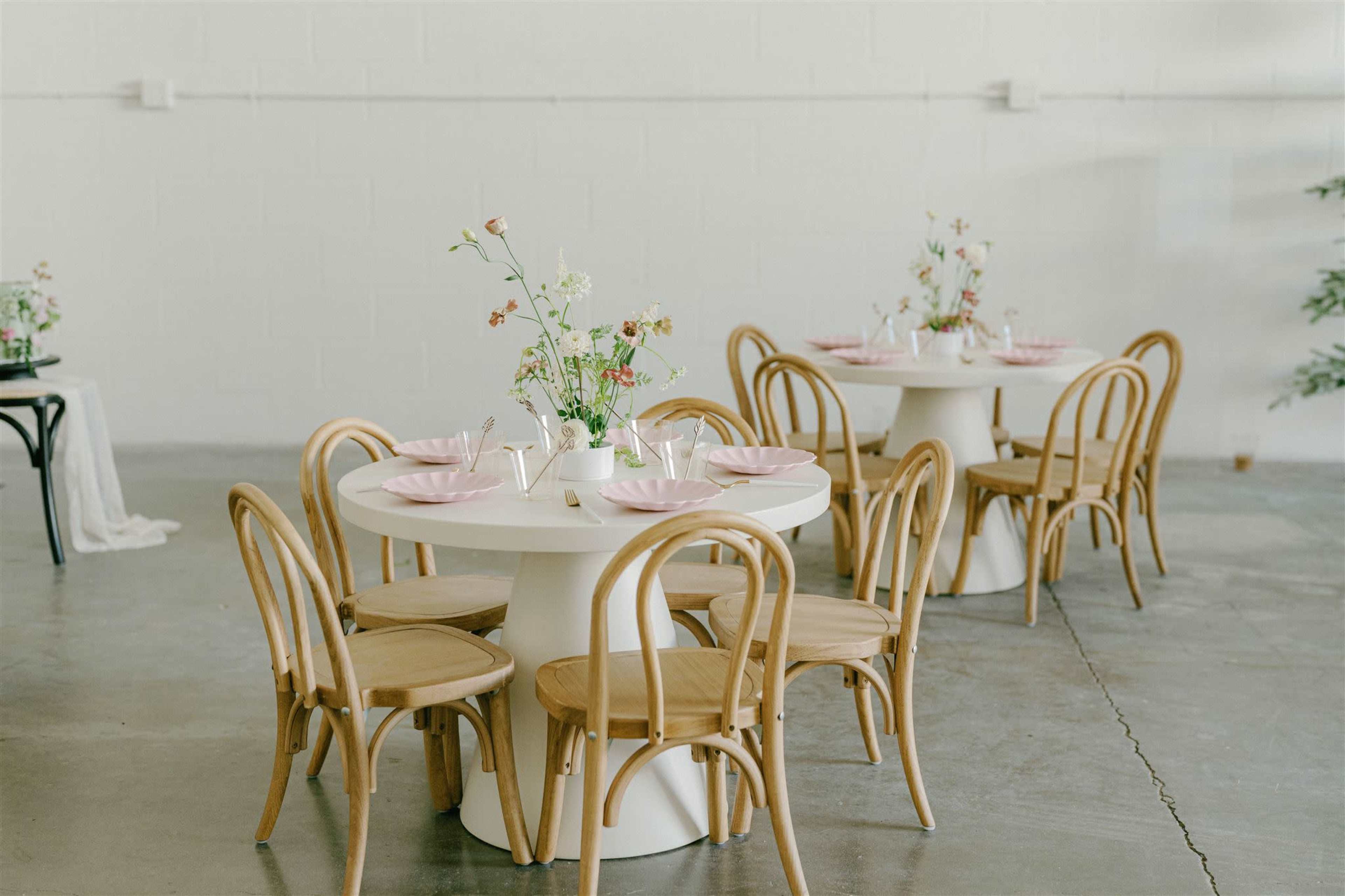 A minimalistic dining setup with round tables, bentwood chairs, and pastel plates, accented by small flower arrangements.