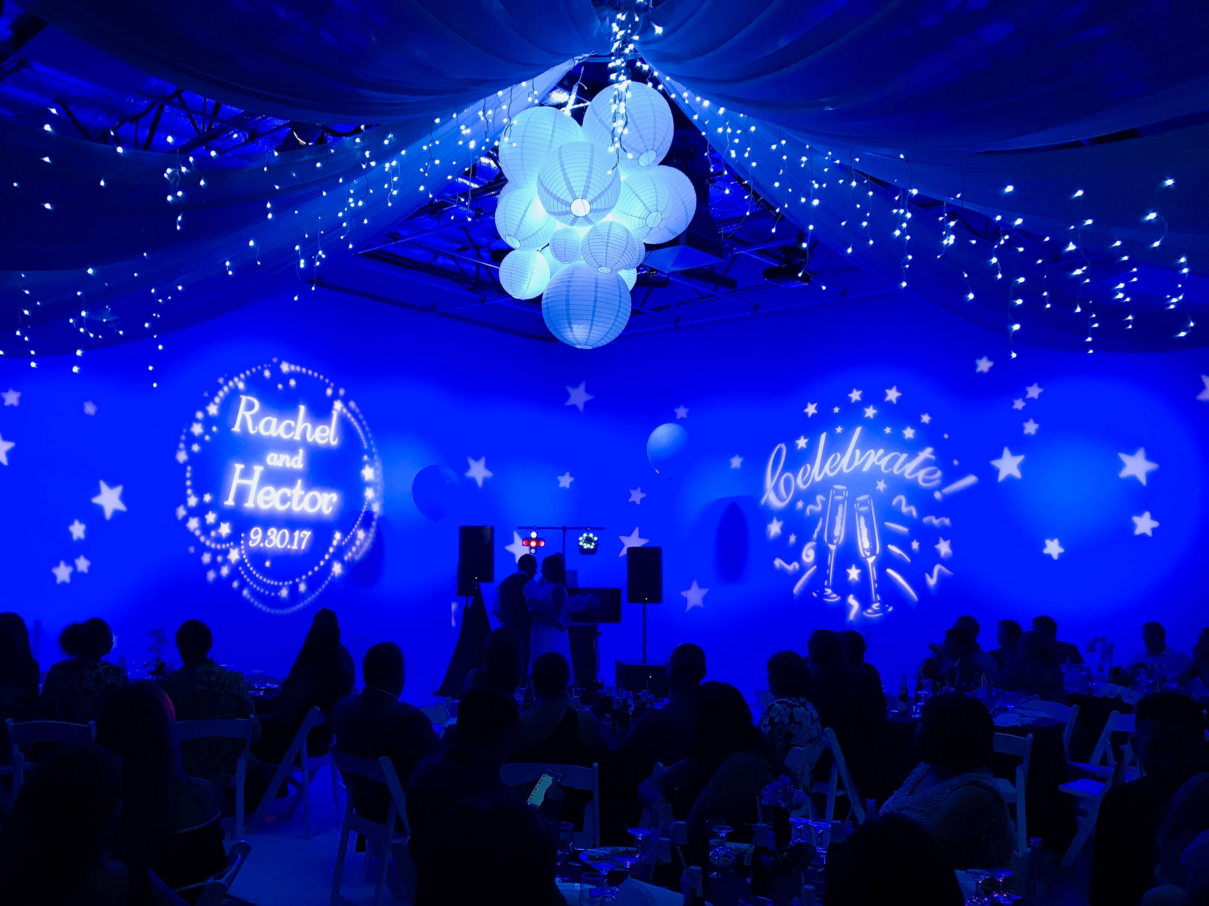 A decorated event space with blue lighting, white hanging lanterns, and projections of "Rachel and Hector" and "Celebrate" on the walls, while guests sit at tables.