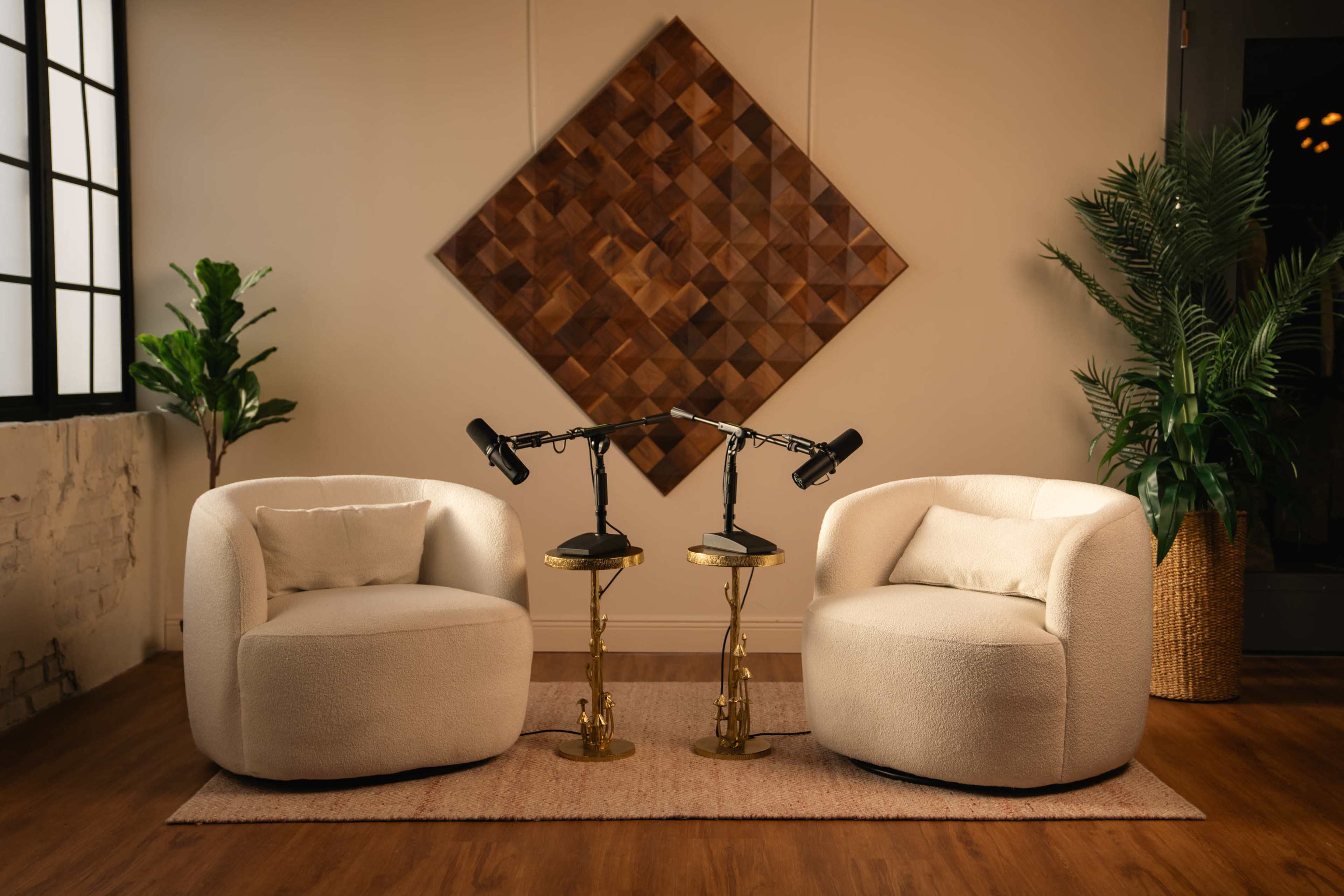The image features two rounded, white upholstered chairs facing each other, accompanied by two gold side tables with black lamps, against a backdrop of a large, wooden geometric wall panel and a potted plant.