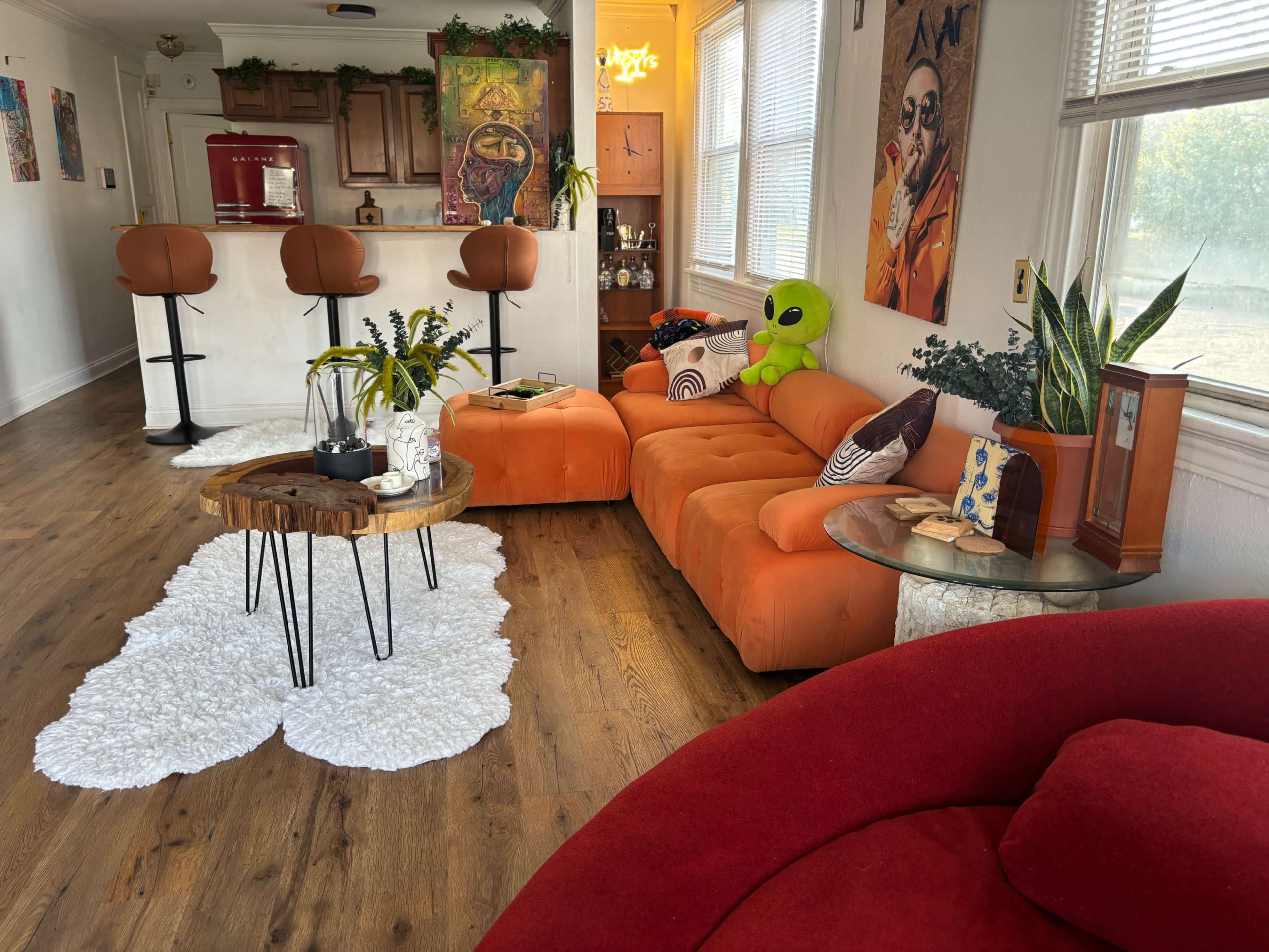 The room features a mix of vibrant orange and red furniture, with a round wooden coffee table on a white rug and a bar area in the background.