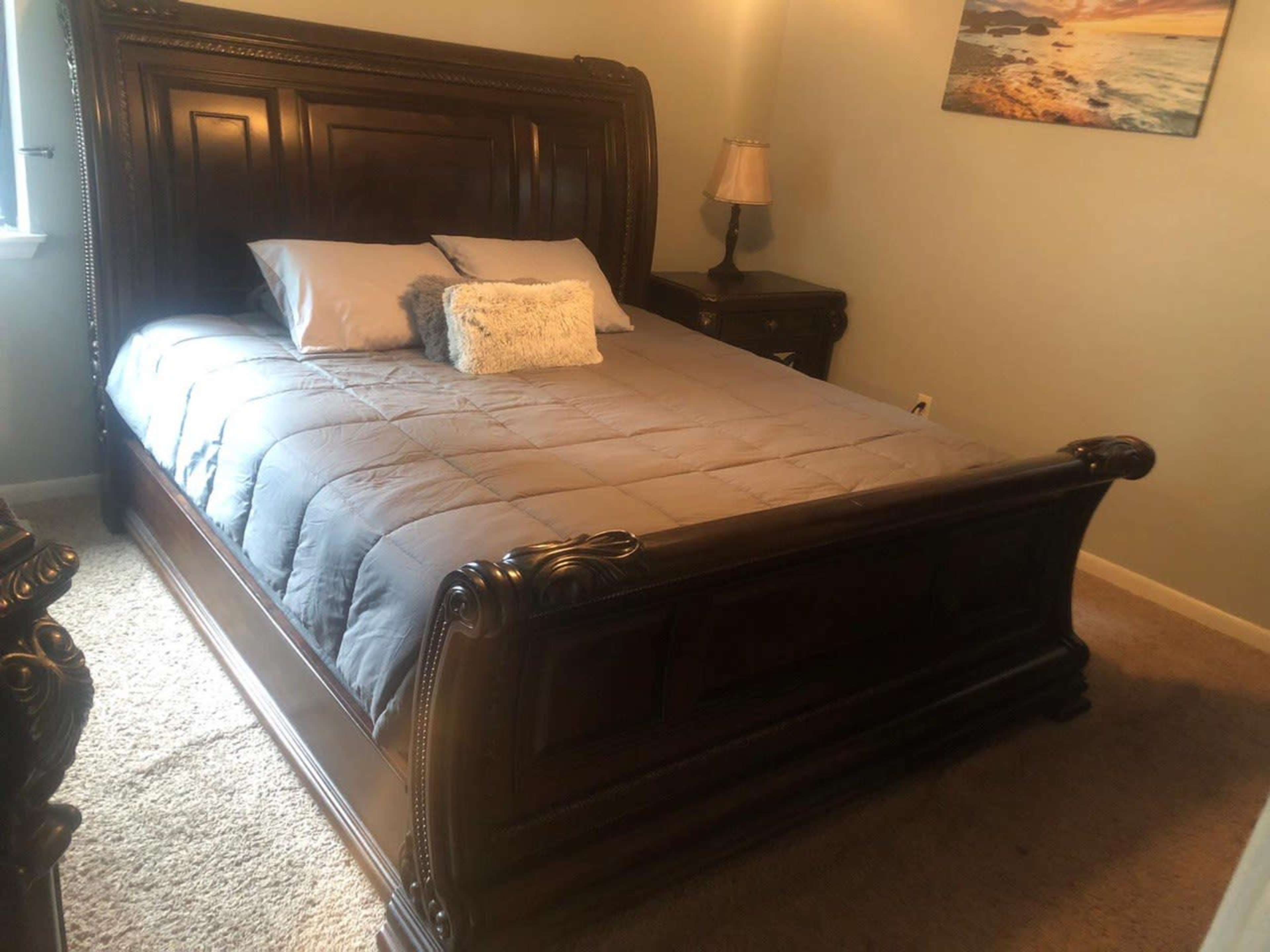 A furnished bedroom featuring a large wooden sleigh bed with gray bedding, a nightstand with a lamp, and a piece of wall art depicting a beach scene.