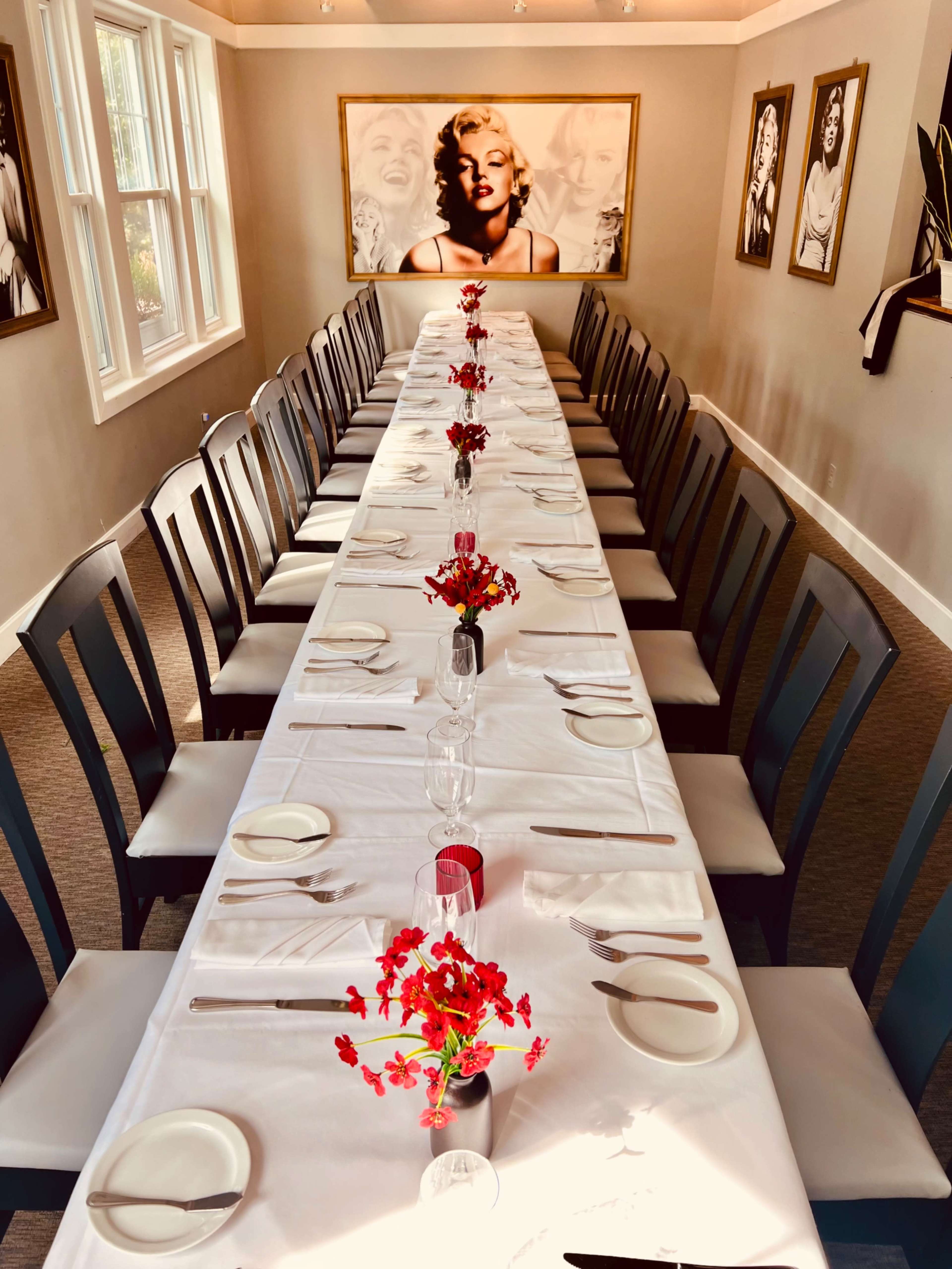 A long, elegantly set dining table with white tablecloths and small floral centerpieces is positioned in a room adorned with black-and-white portraits.