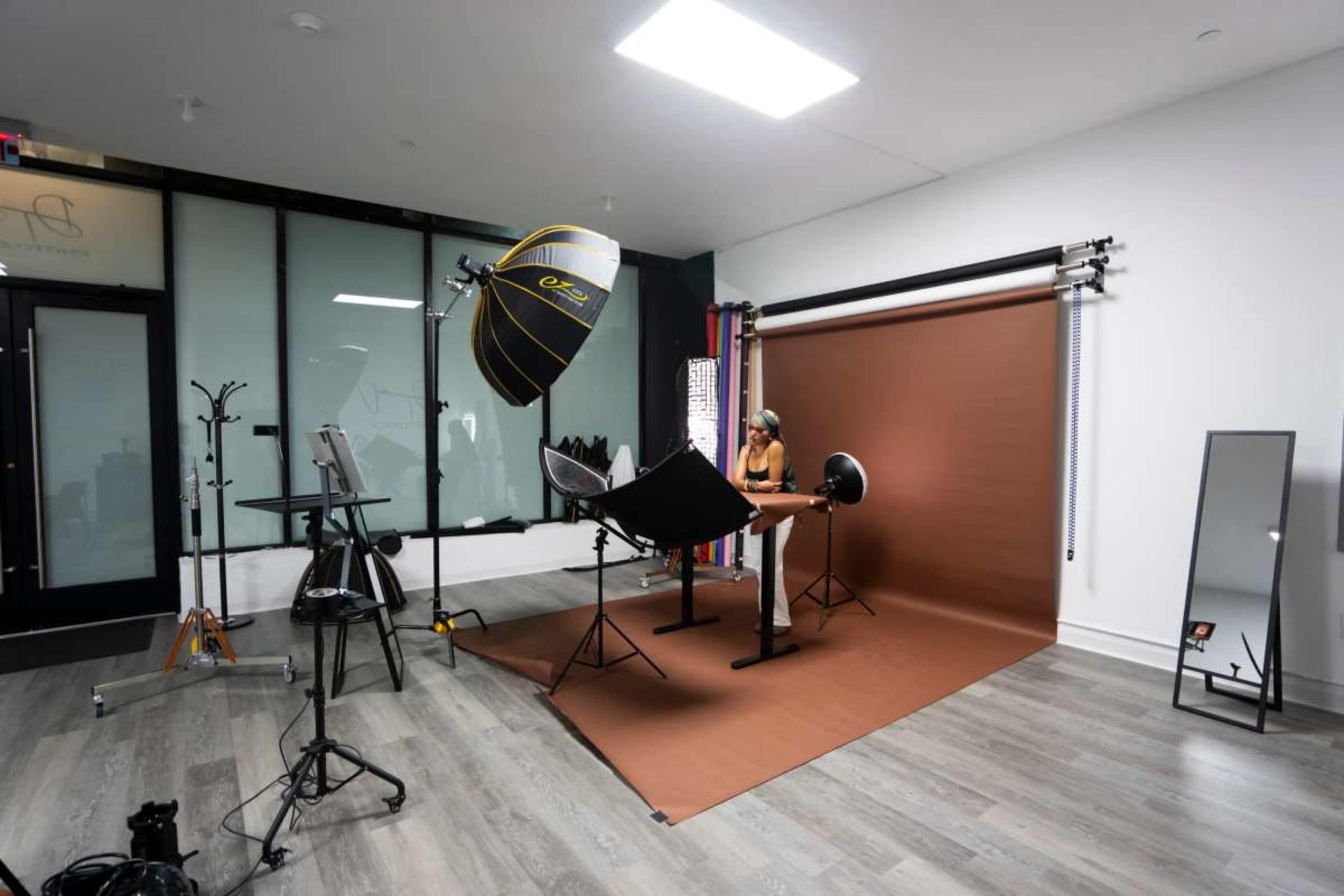 The image shows a photography studio with a model sitting at a table, surrounded by lighting equipment and backdrops.