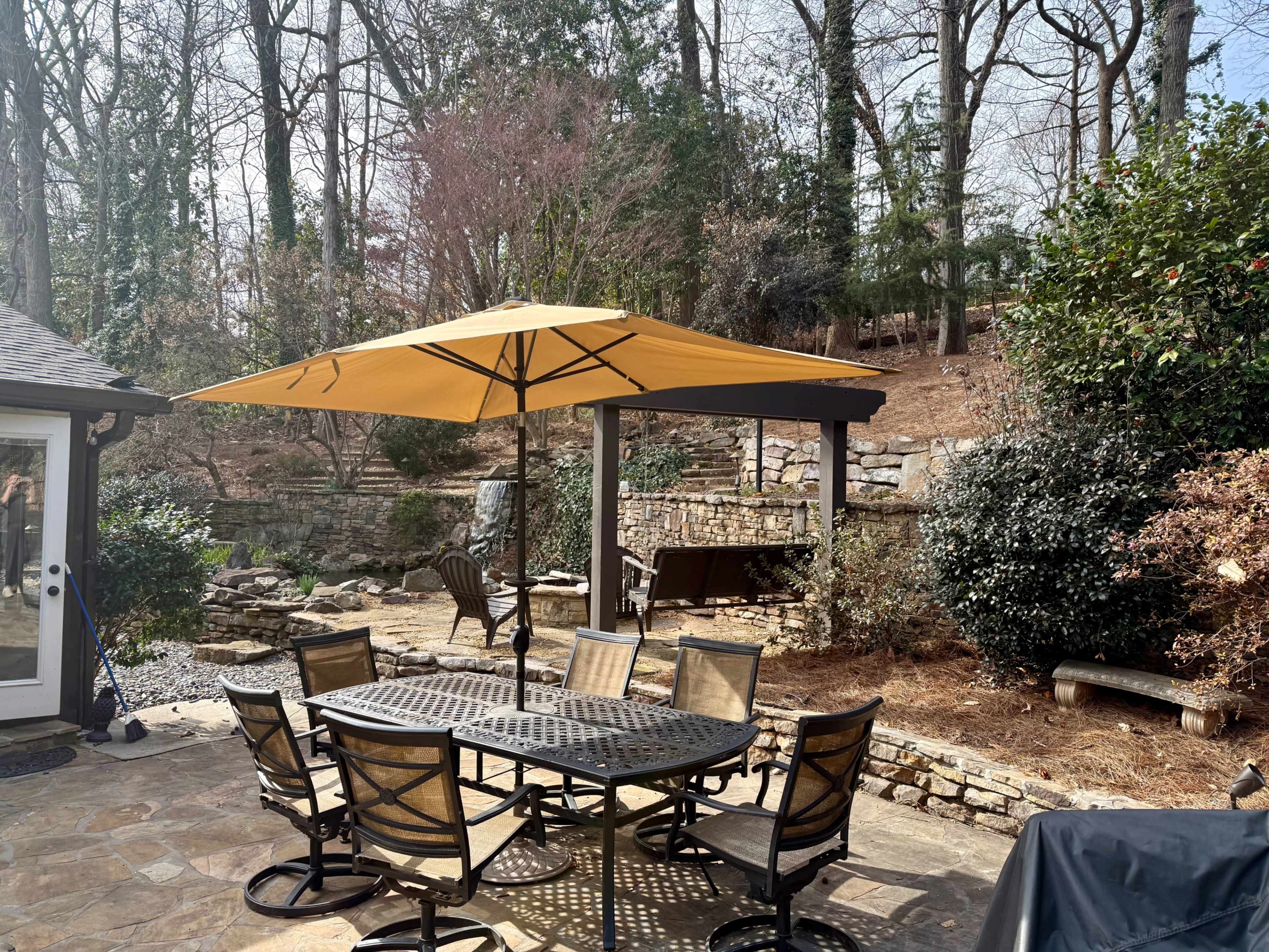 A patio area features a round table surrounded by six chairs and an orange umbrella, with a backdrop of trees and stone landscaping.