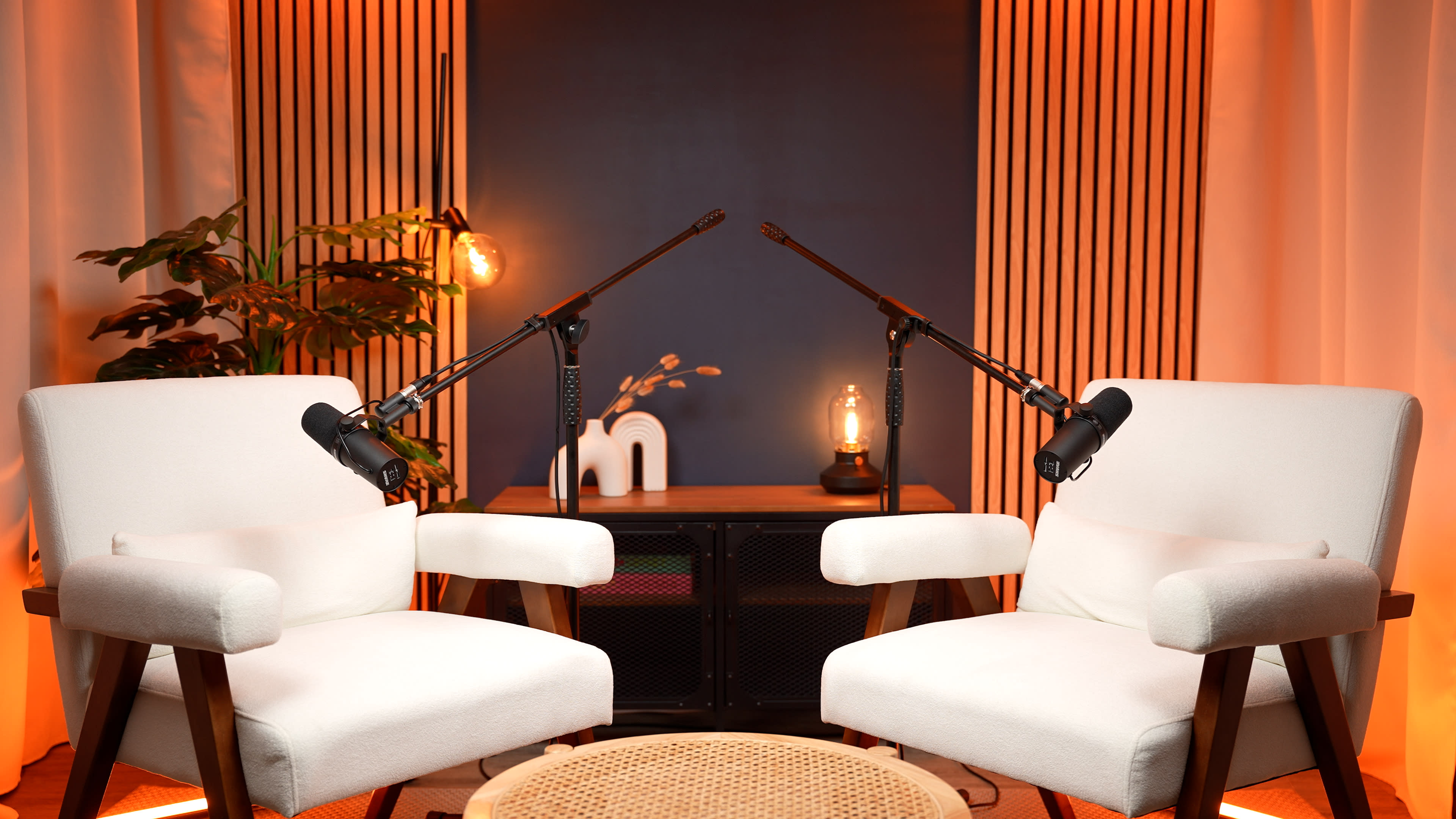 The image shows two white chairs positioned opposite each other, each with a microphone mounted on a boom arm, against a backdrop of orange and neutral-colored decor.