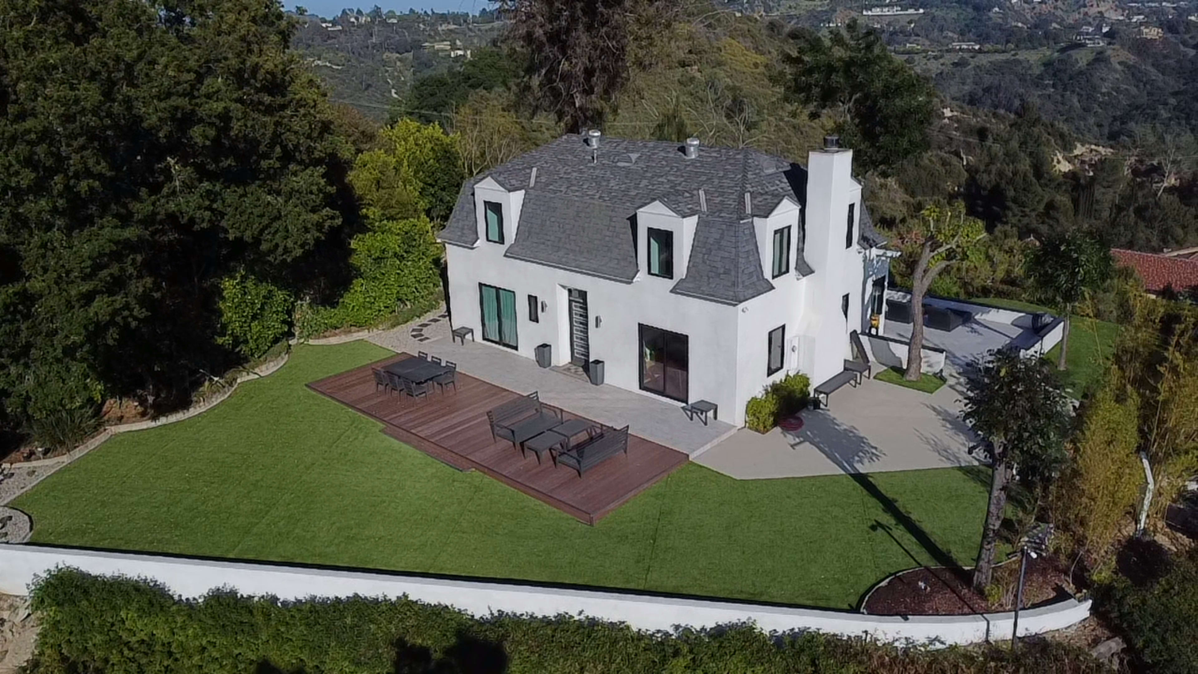 The image shows a two-story white house with a gray shingle roof, surrounded by a manicured lawn and featuring a wooden deck with outdoor furniture.