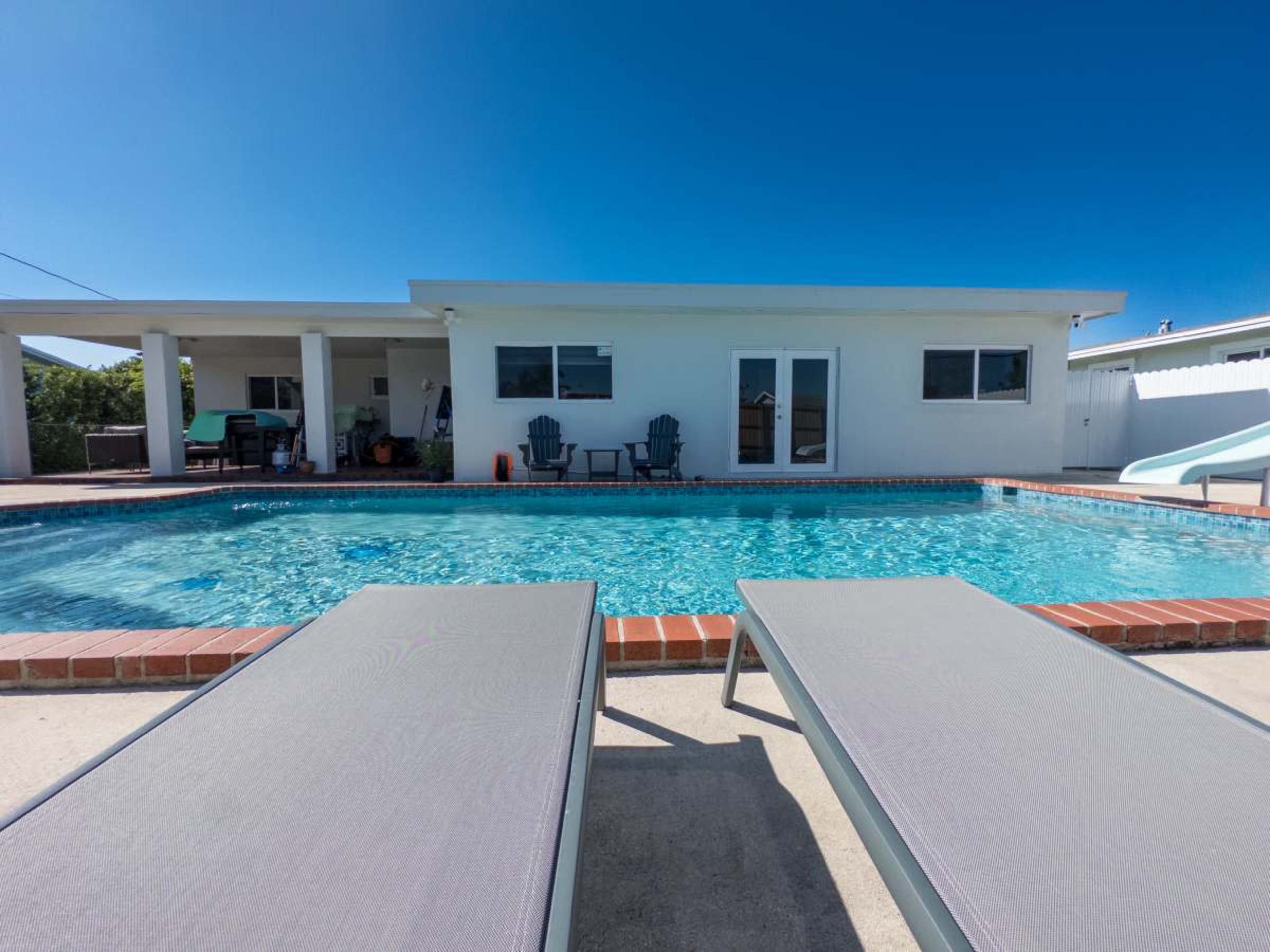 A clear swimming pool is surrounded by lounge chairs and a house with sliding glass doors in the background.