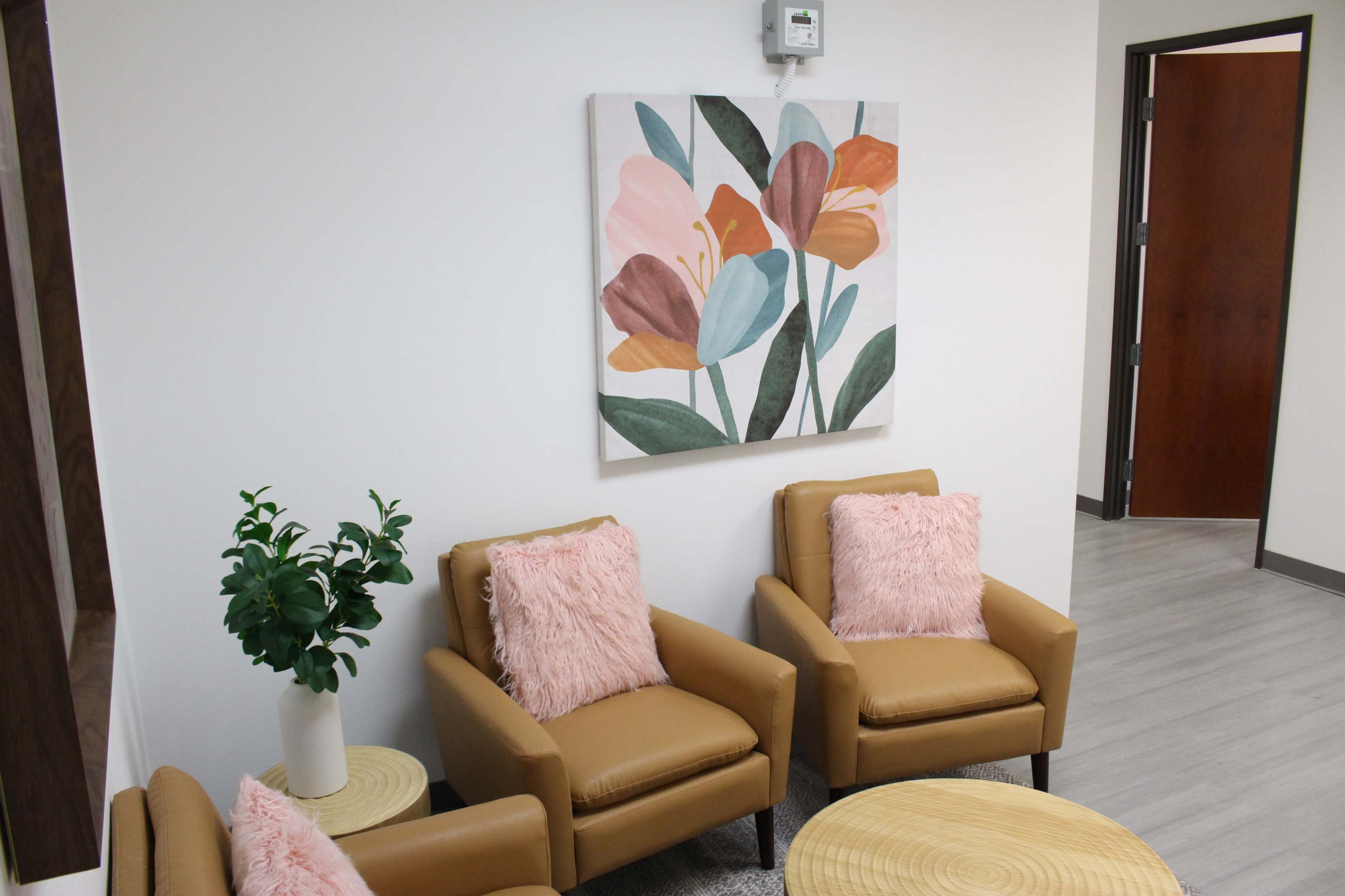 A waiting area with two tan armchairs, each adorned with a pink cushion, a round woven table, and a floral artwork on the wall.