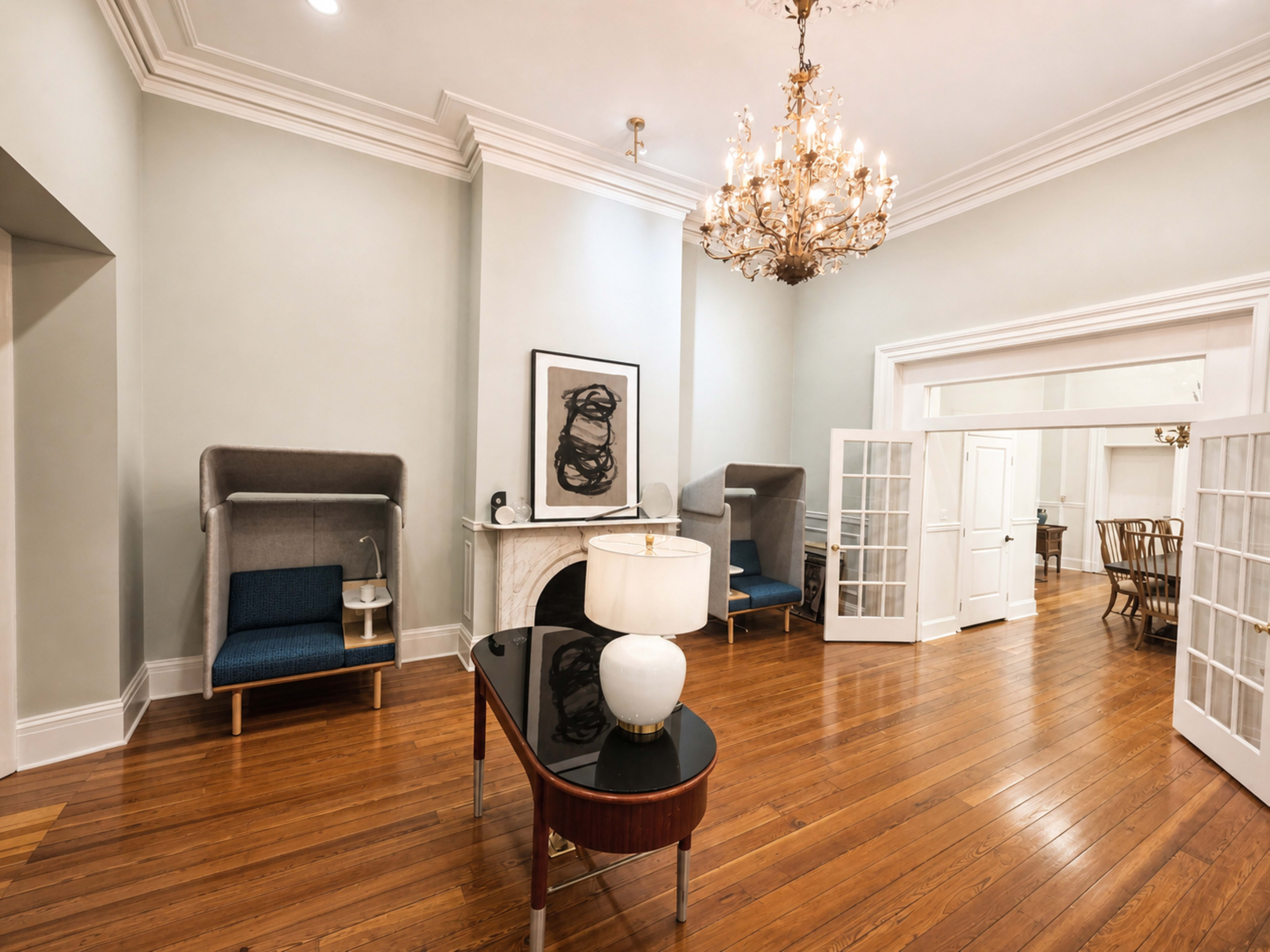 The image shows a spacious room with hardwood floors, featuring a chandelier, a modern lamp on a coffee table, and two blue upholstered chairs flanking a fireplace.