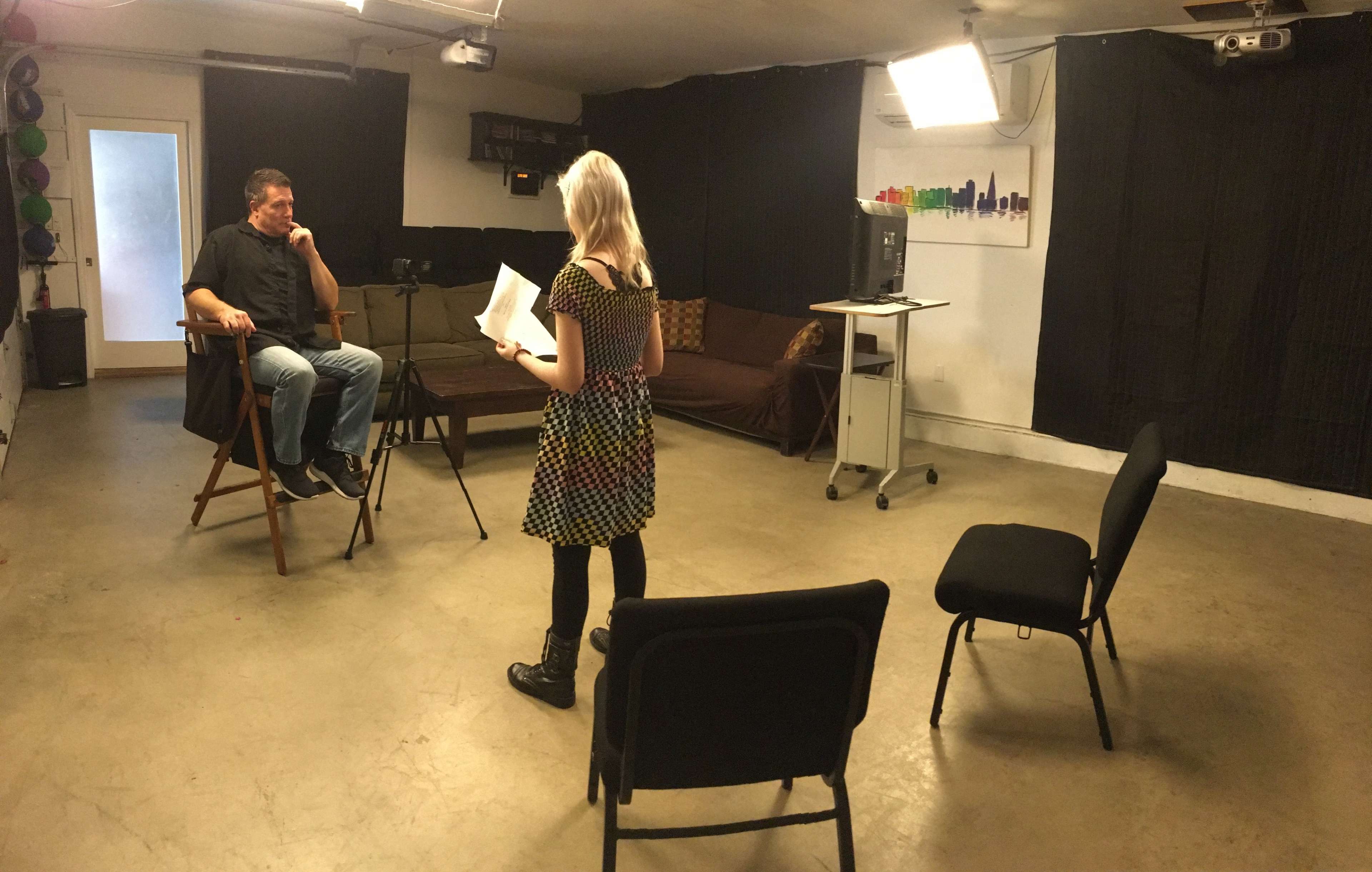 A woman stands facing a seated man in a film studio, holding a script, with cameras and chairs arranged in the background.