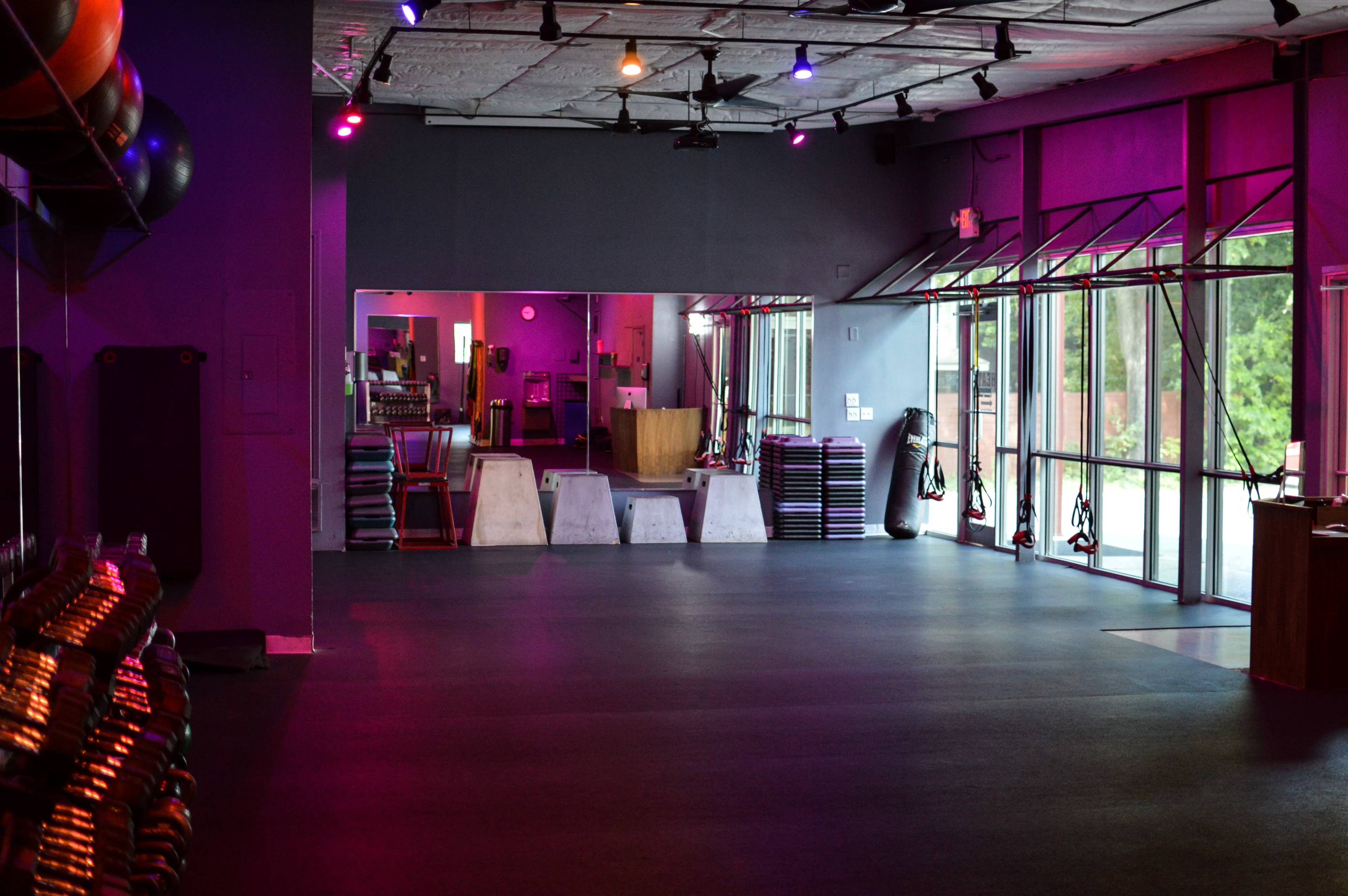 An empty gym interior with purple lighting, exercise equipment stacked against the walls, and large windows allowing natural light to enter.