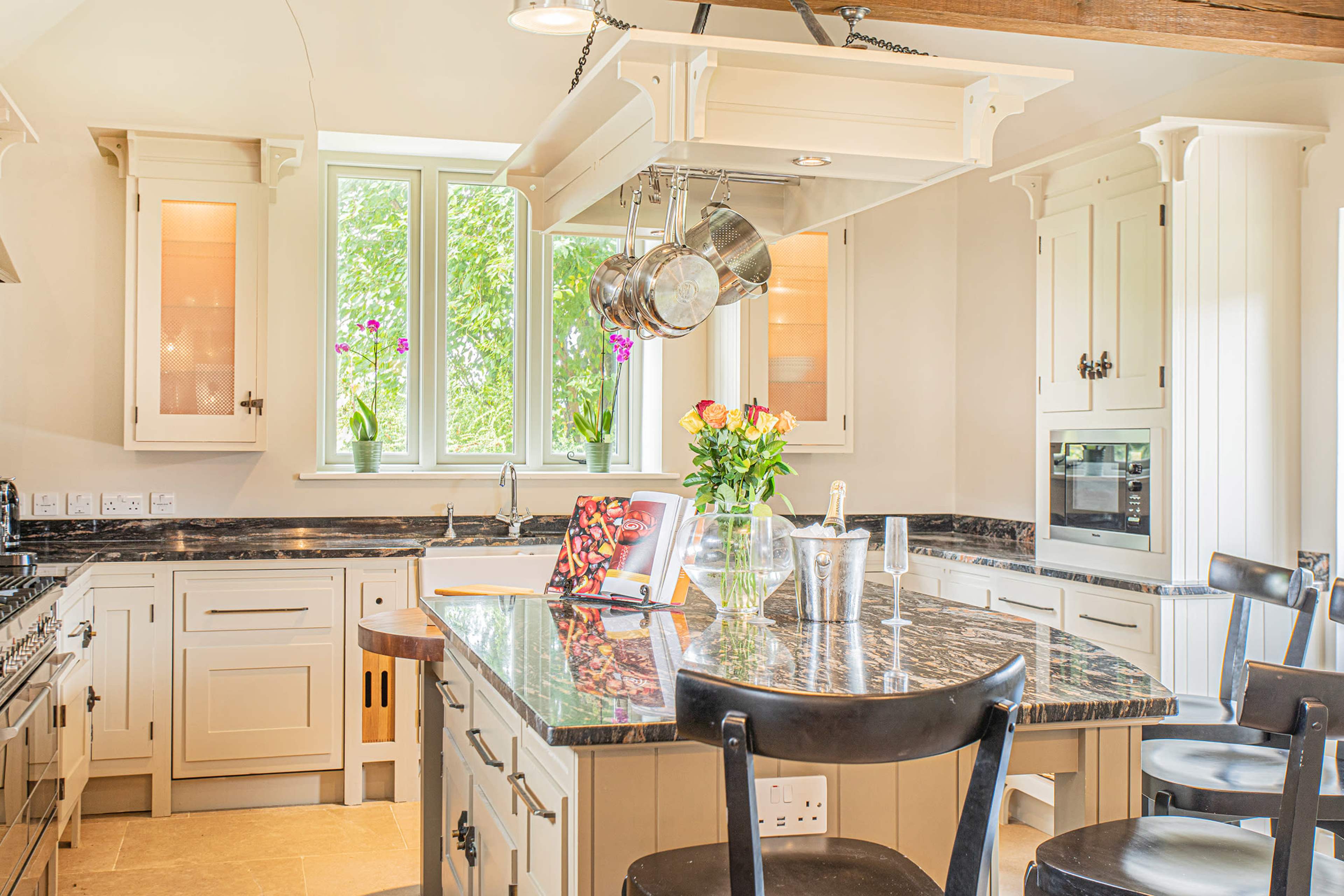 The kitchen features a central island with a marble countertop, surrounded by black chairs, and includes windows that provide natural light and view of greenery outside.