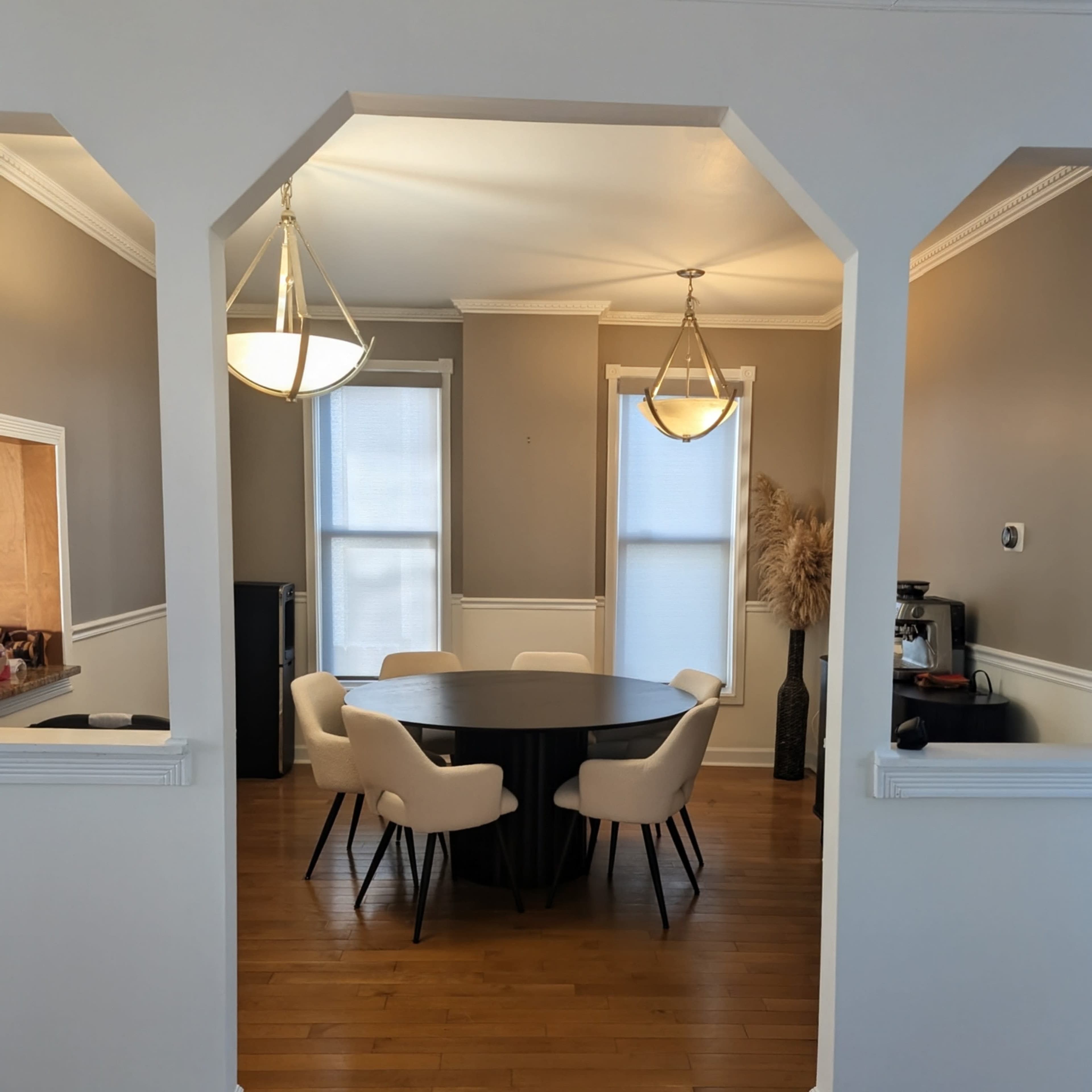 The image shows a dining area with a round table surrounded by six white chairs, illuminated by two pendant lights, and featuring large windows and light-colored walls.