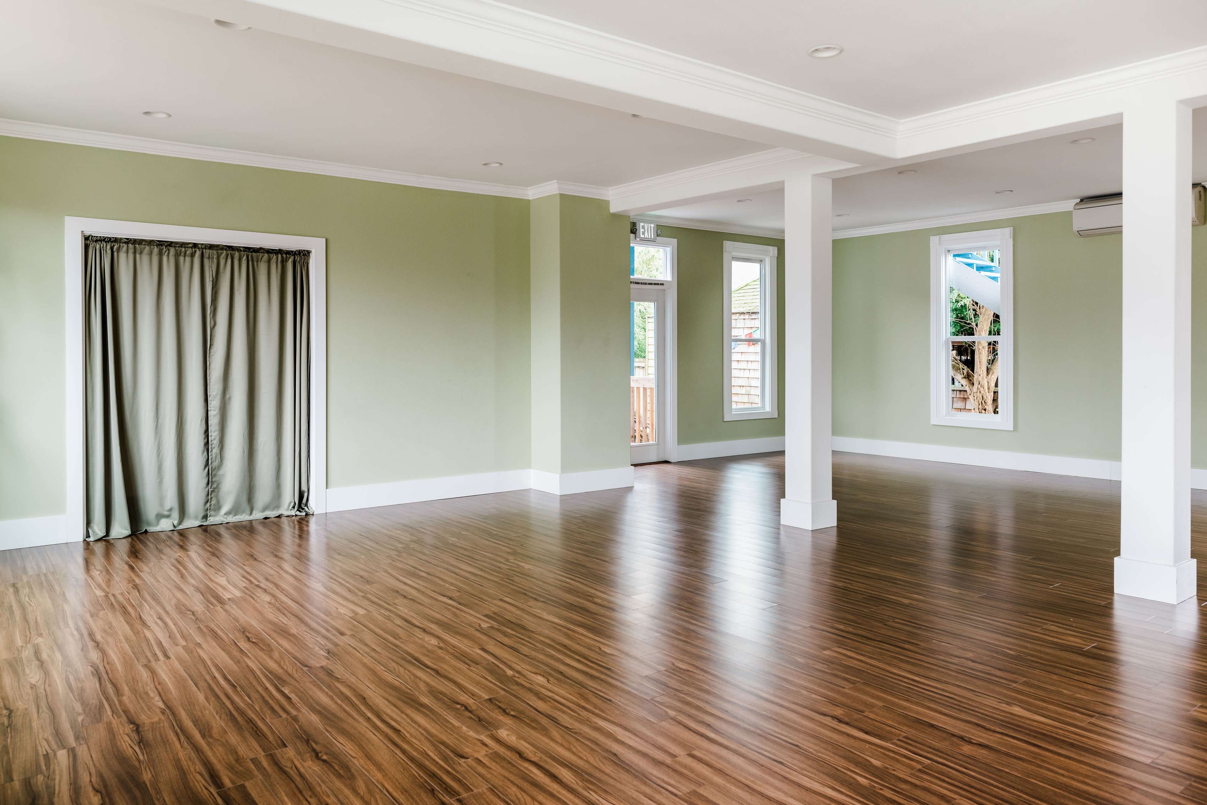 The image shows a spacious, empty room with green walls, hardwood flooring, and large windows.