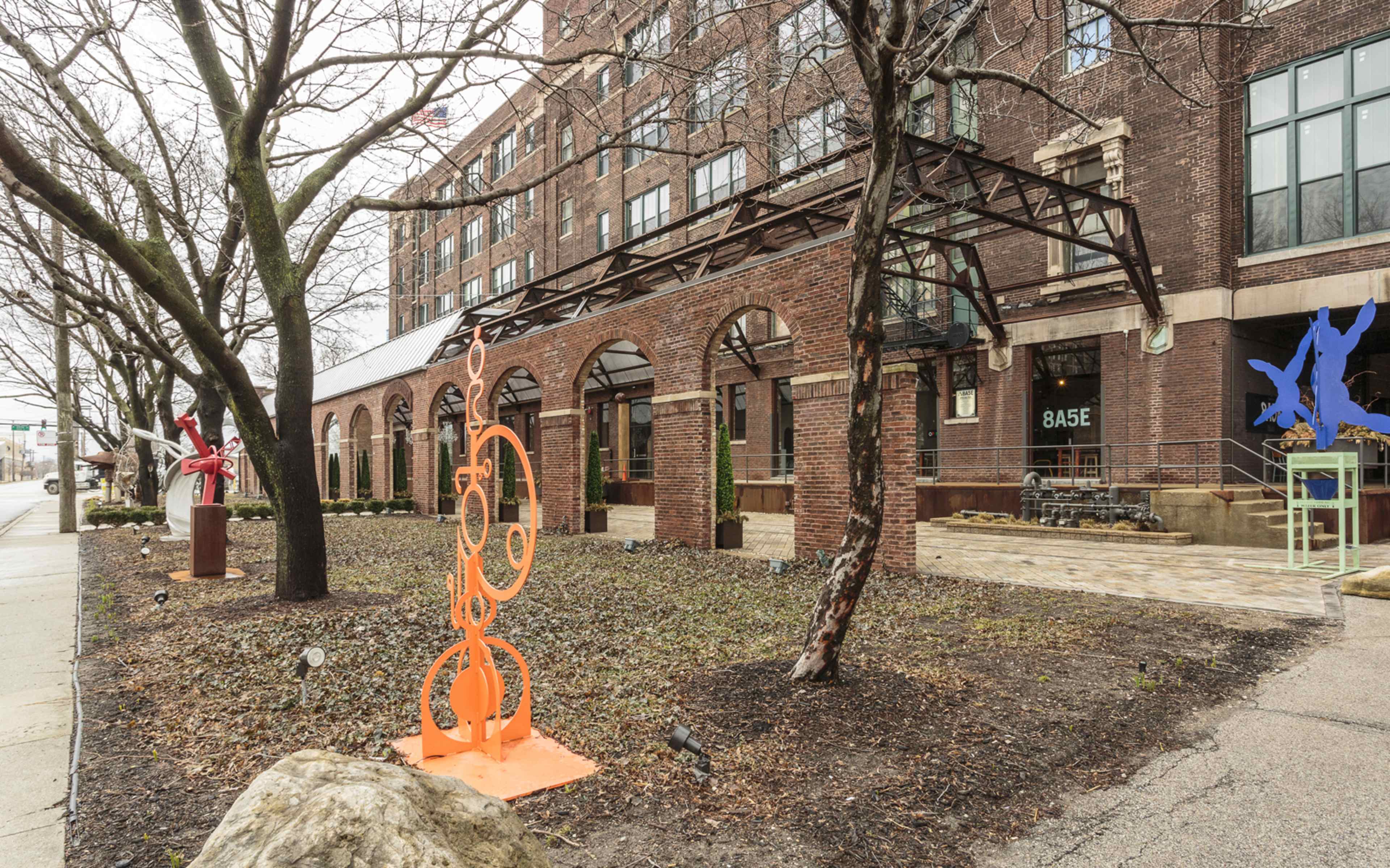 The image shows a brick building with large windows, surrounded by trees and featuring colorful sculptures in a landscaped area.