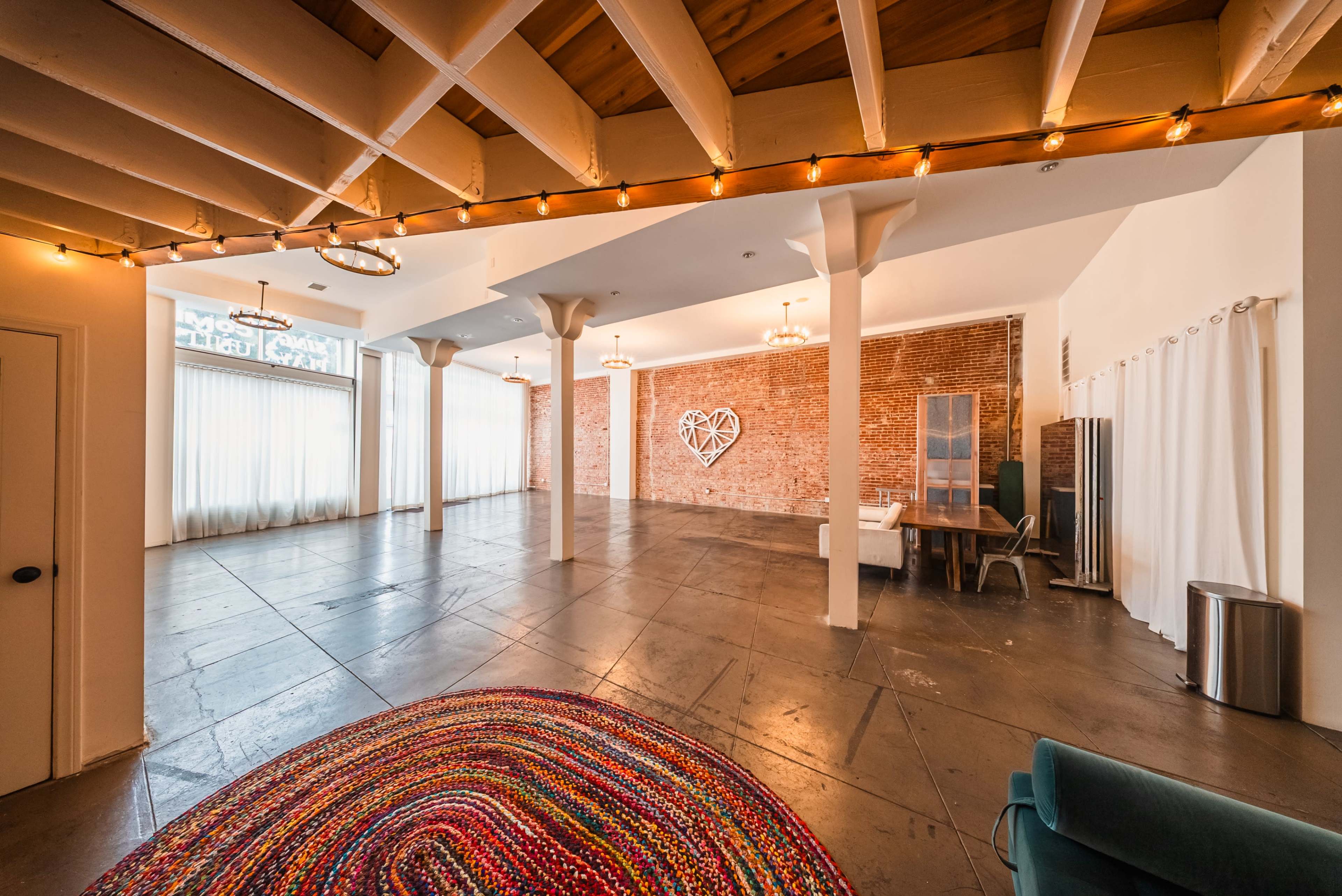 The image depicts a spacious, well-lit room featuring exposed brick walls, string lights, and a circular woven rug on the concrete floor.