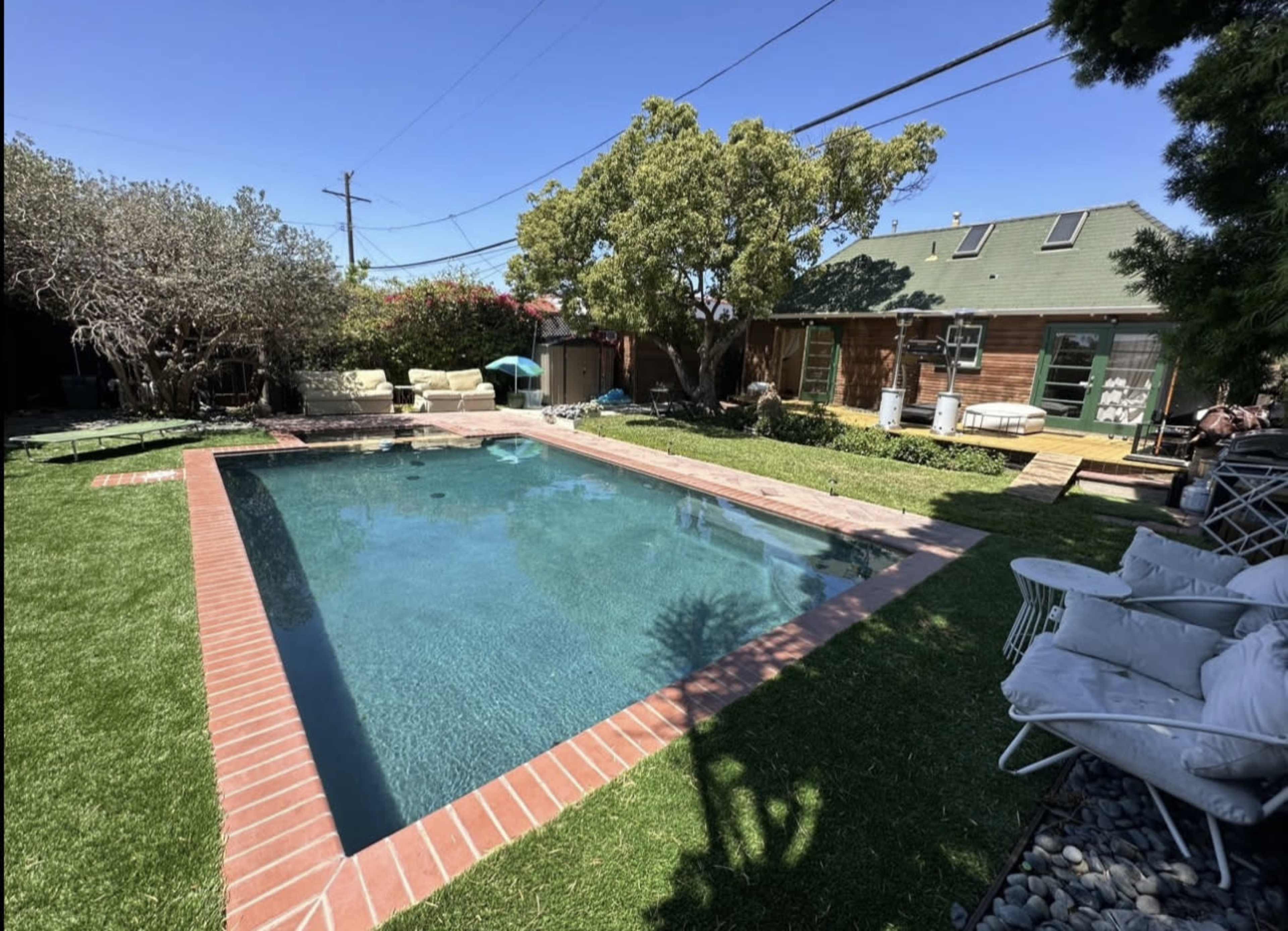 A backyard featuring a rectangular swimming pool surrounded by a brick border, green grass, and some seating areas under trees.