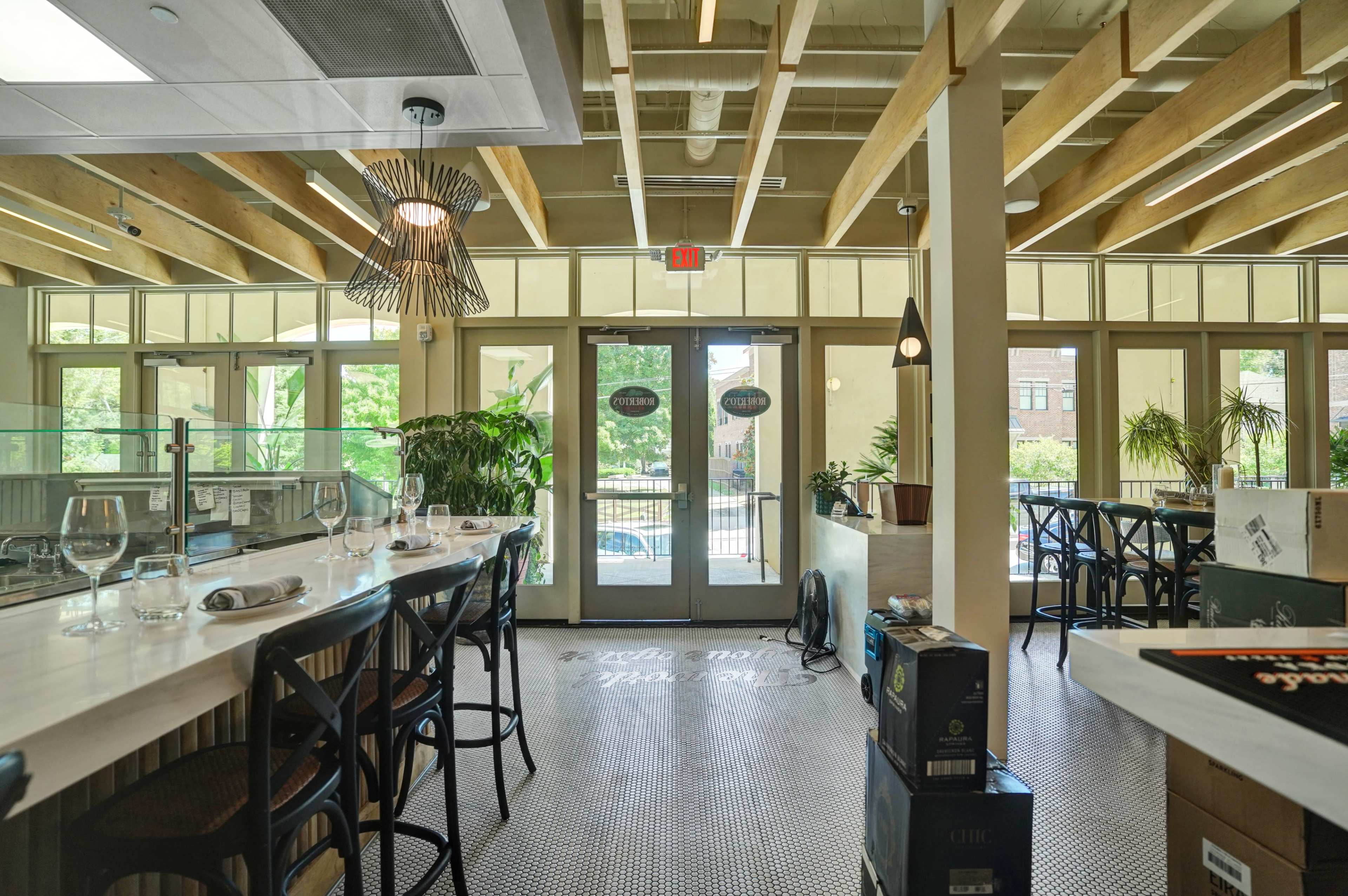 The image shows the interior of a modern restaurant featuring a long bar area with high stools, large windows inviting natural light, and plants decorating the space.