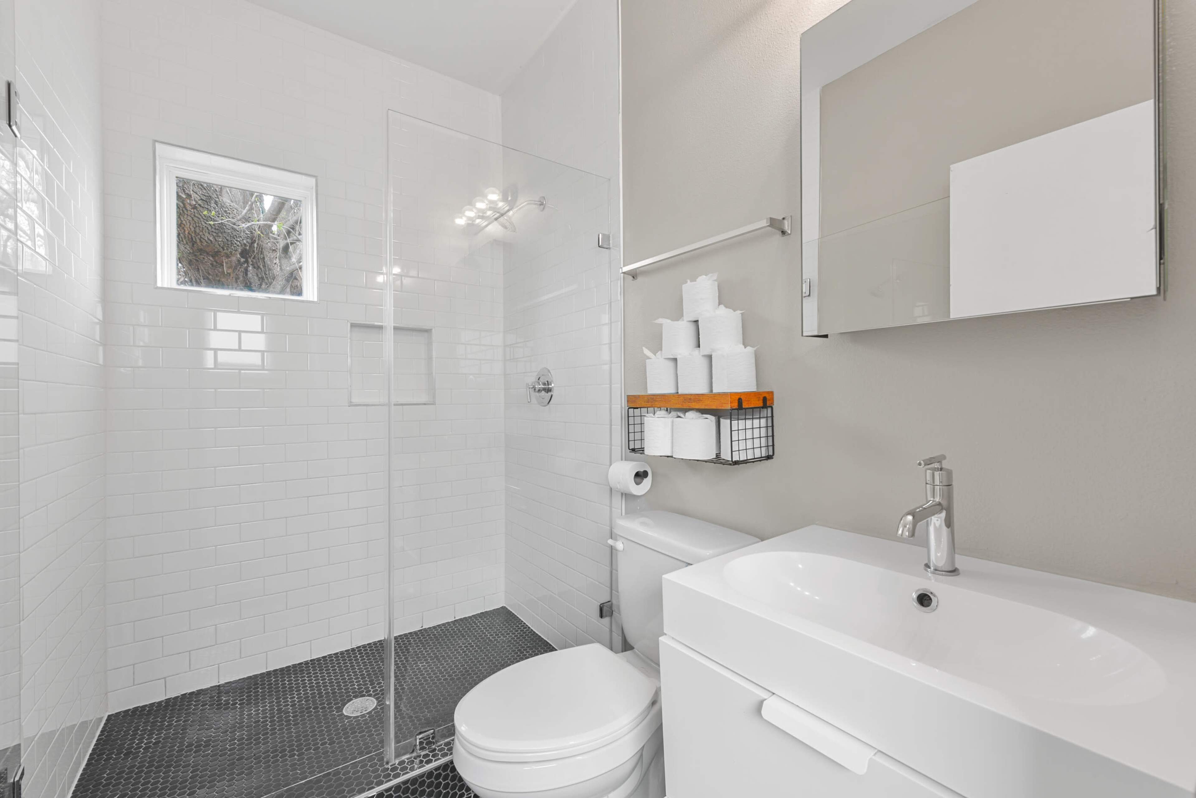 The image shows a modern bathroom with a glass walk-in shower, white tiles, a toilet, and a sink with a mirror above it.