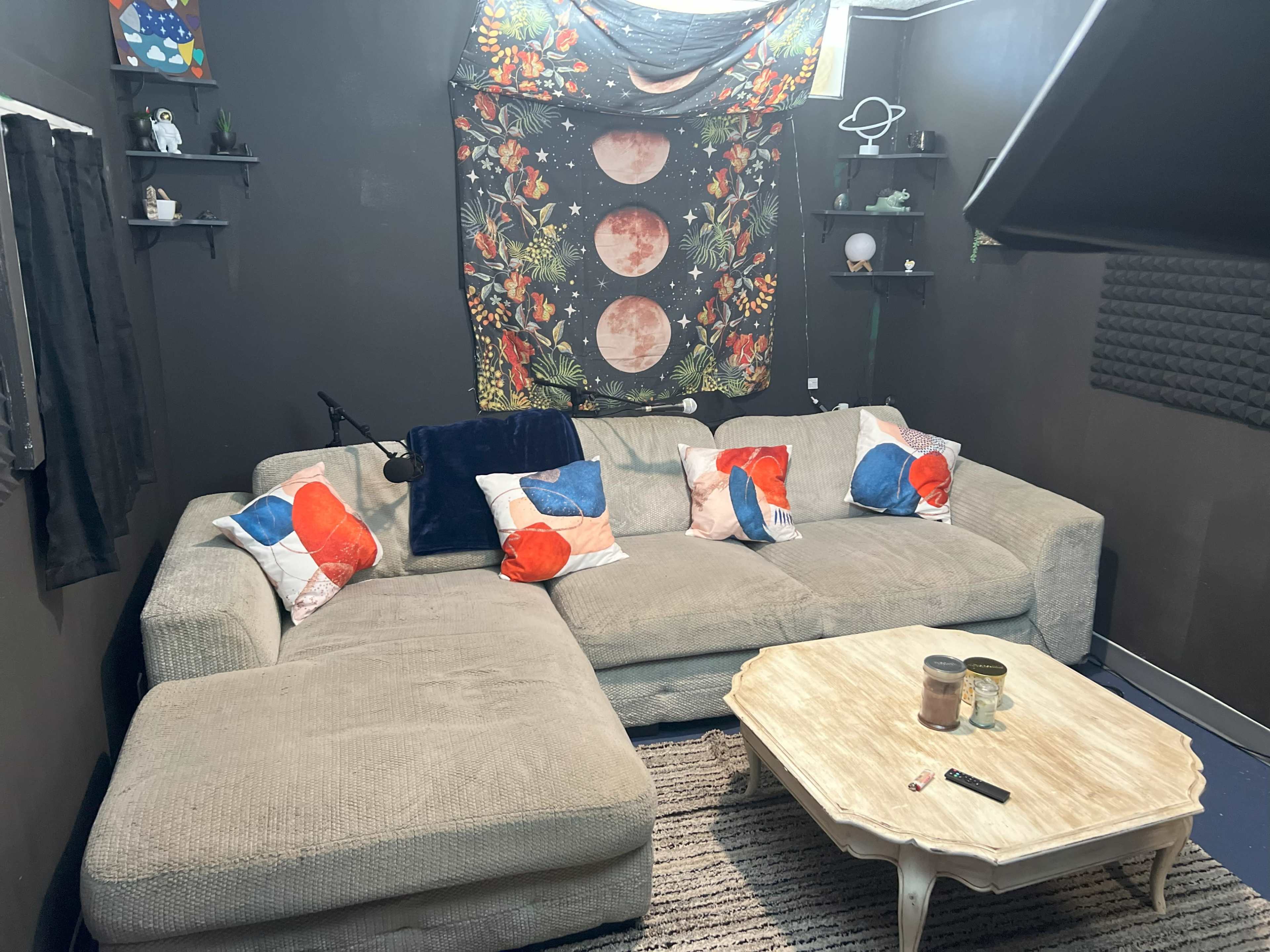 A cozy living room setup featuring a large sectional couch with colorful throw pillows, a small coffee table, and a wall adorned with a tapestry and shelves.
