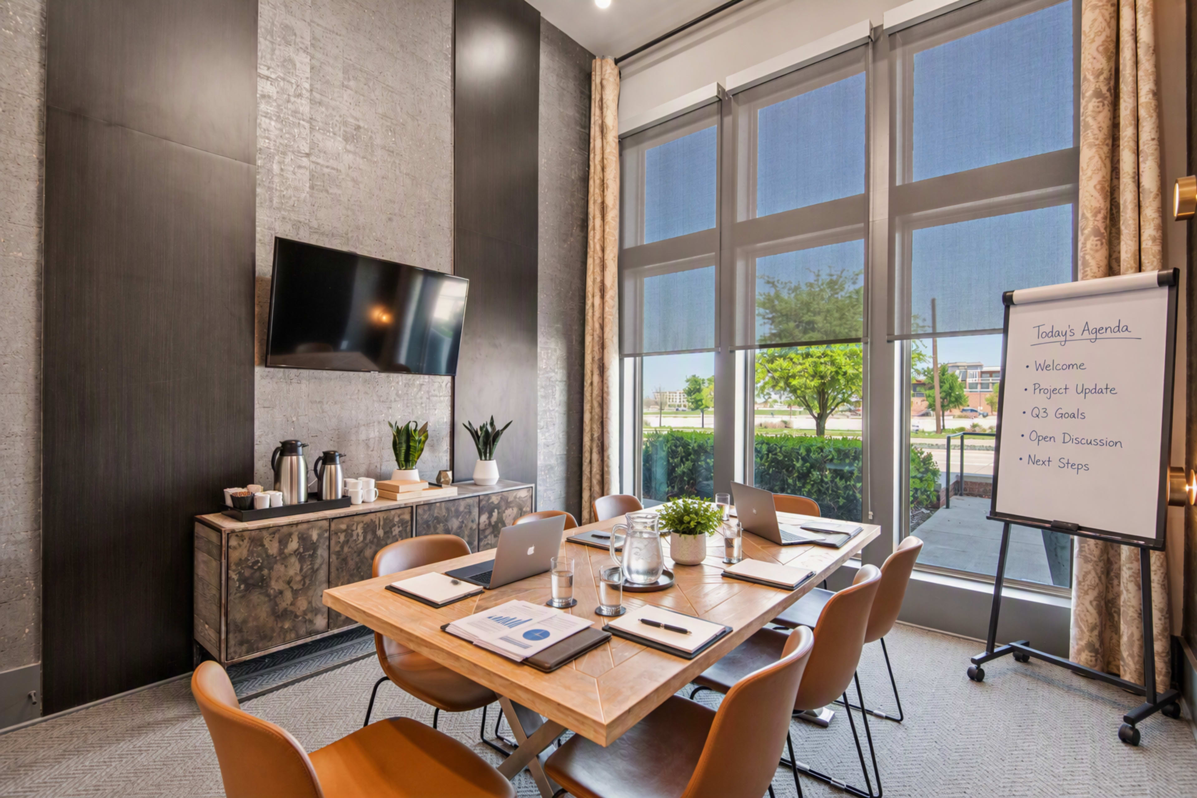 A modern conference room features a wooden table with laptops and notepads, a large screen on the wall, and windows revealing a view of trees outside.