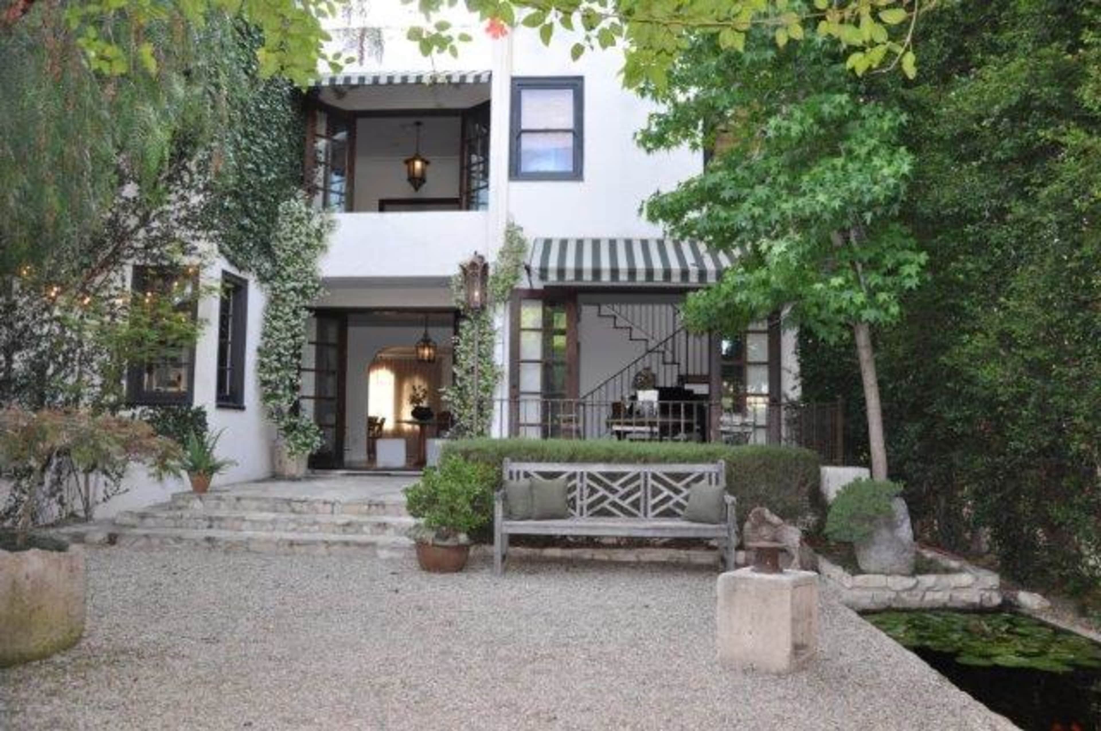 A quaint courtyard with a stone pathway leading to a white house surrounded by greenery and a small pond.