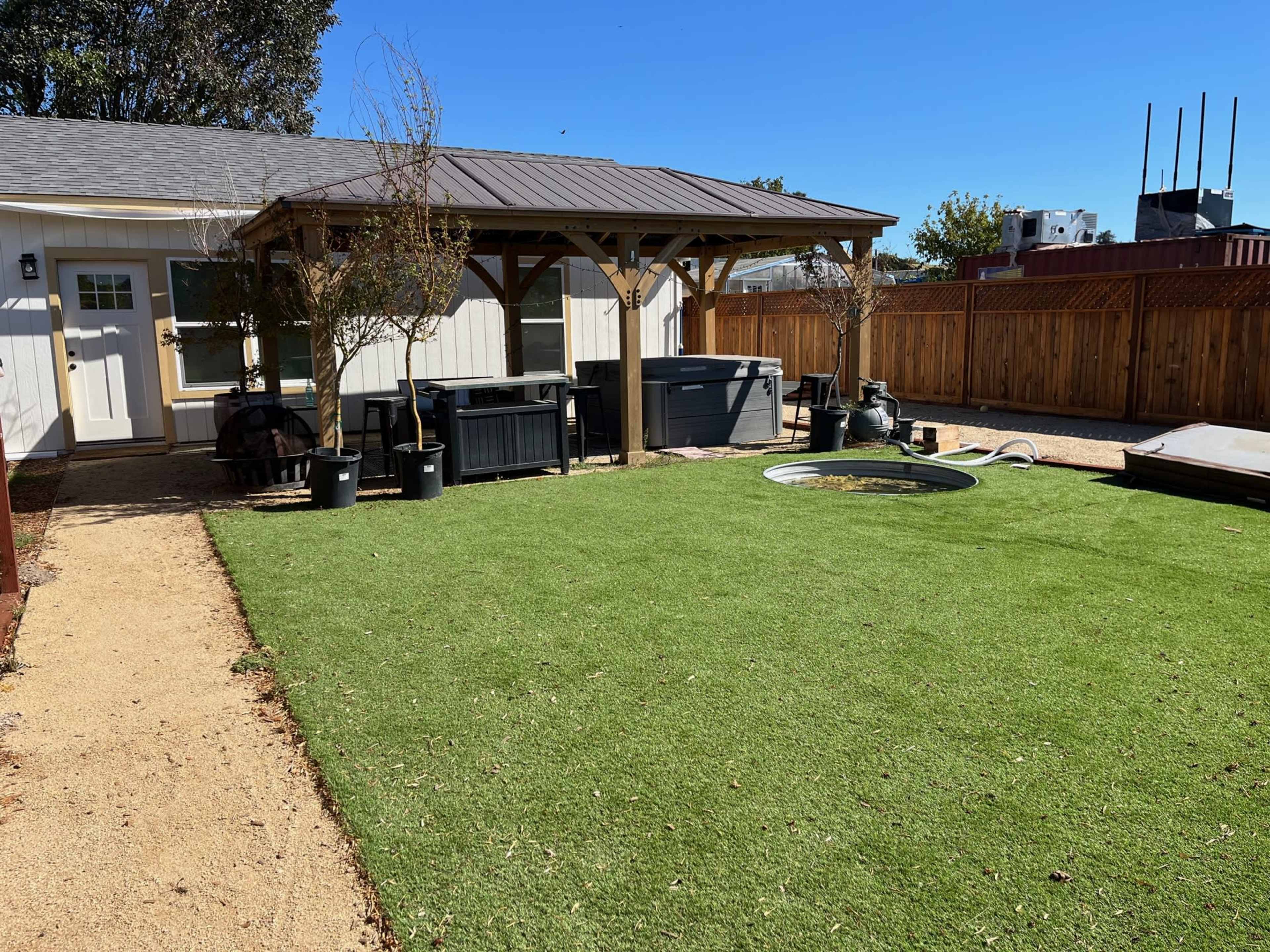 A backyard with a covered patio, a grassy area, hot tubs, and a circular pool surrounded by planters and a wooden fence.