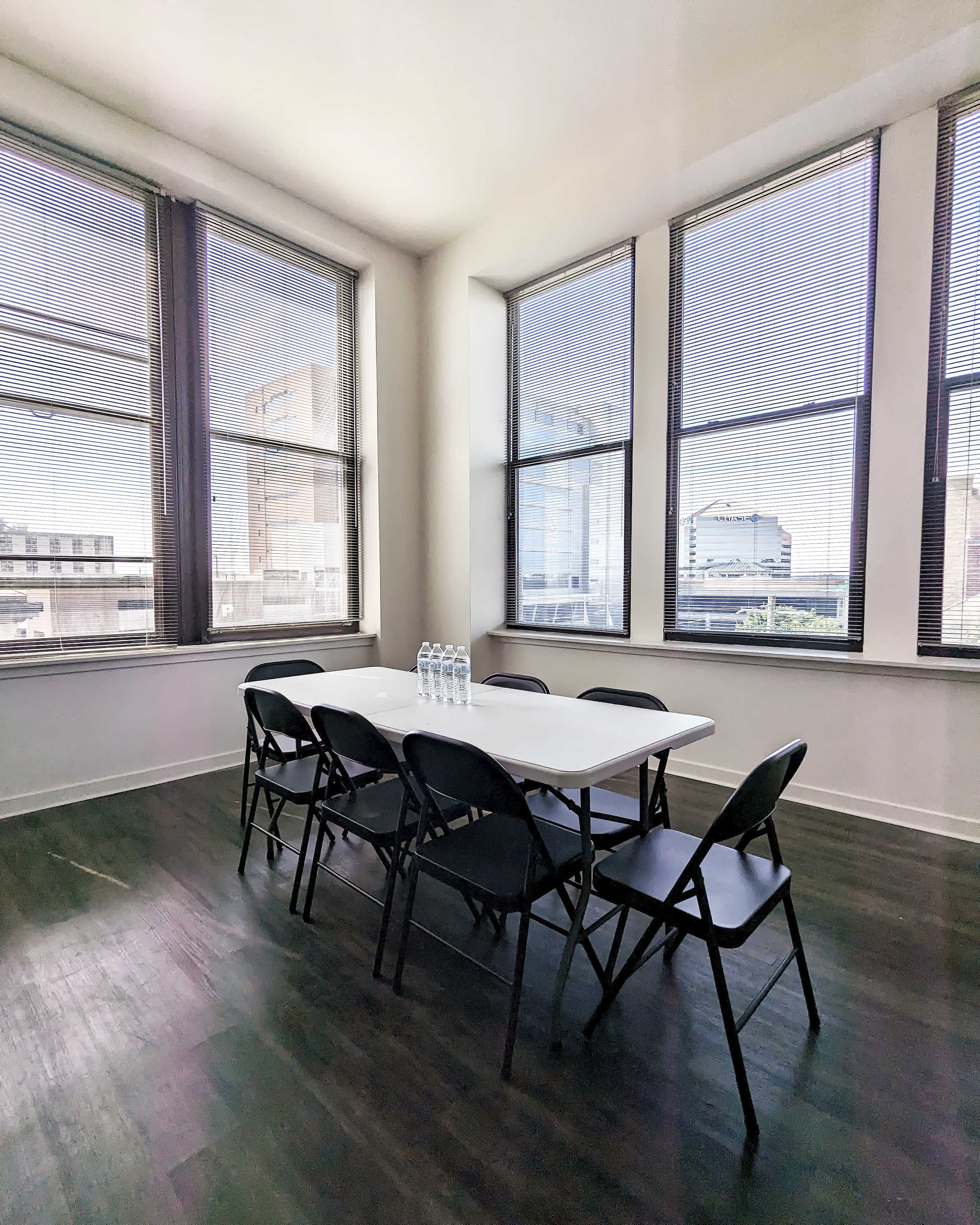 A bright meeting room features a white table surrounded by black chairs, with large windows allowing natural light to enter.