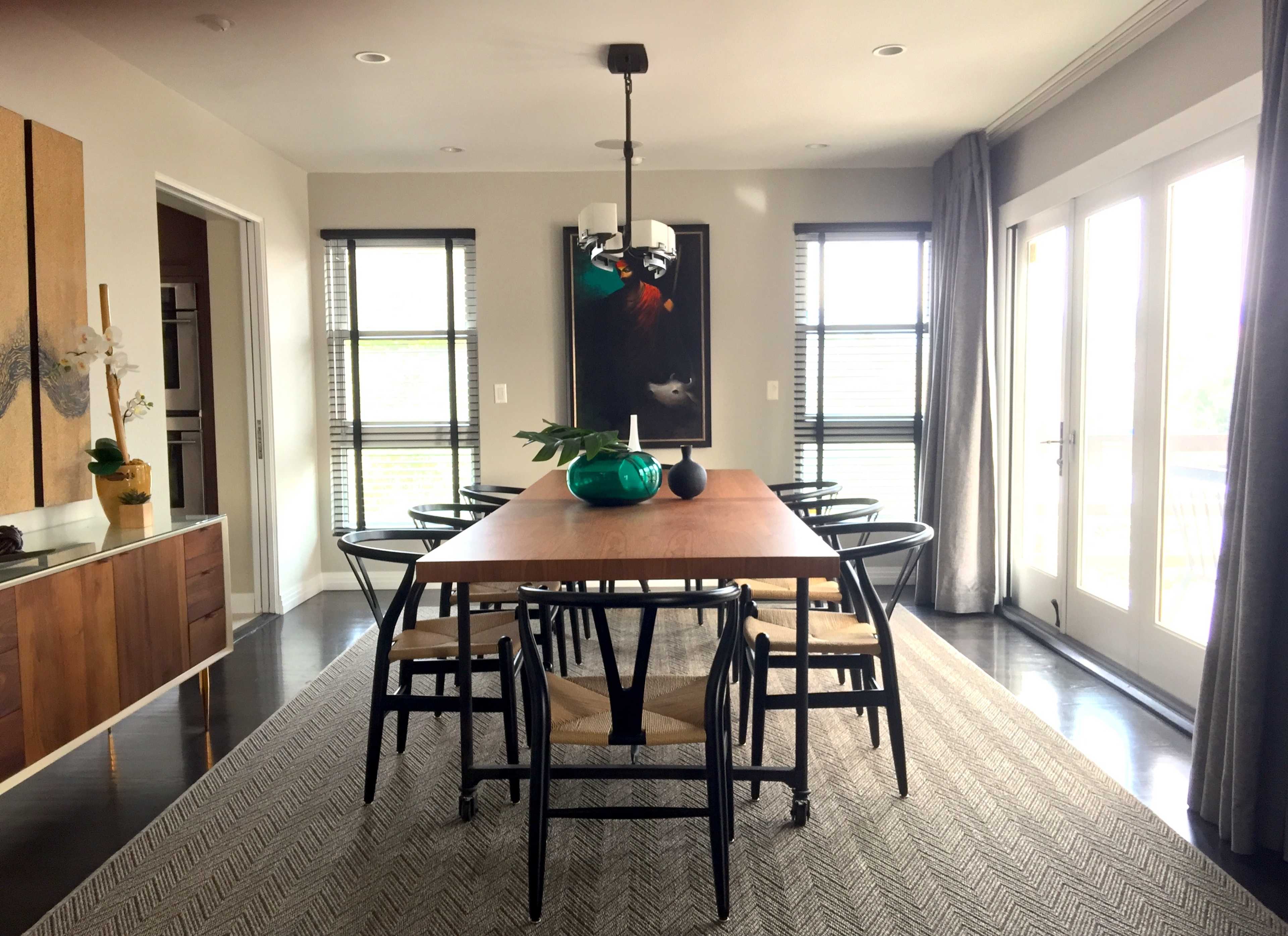 A modern dining room features a long wooden table with black chairs, framed by large windows and a piece of artwork on the wall.