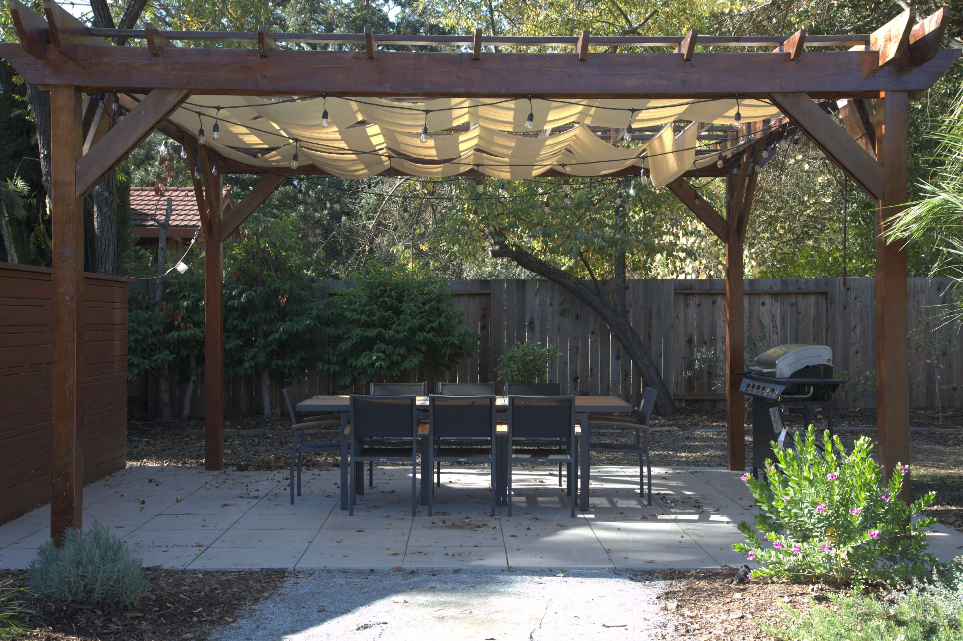 A wooden pergola with a fabric canopy covers a dining table and chairs, situated in a backyard with landscaped borders and a grill nearby.