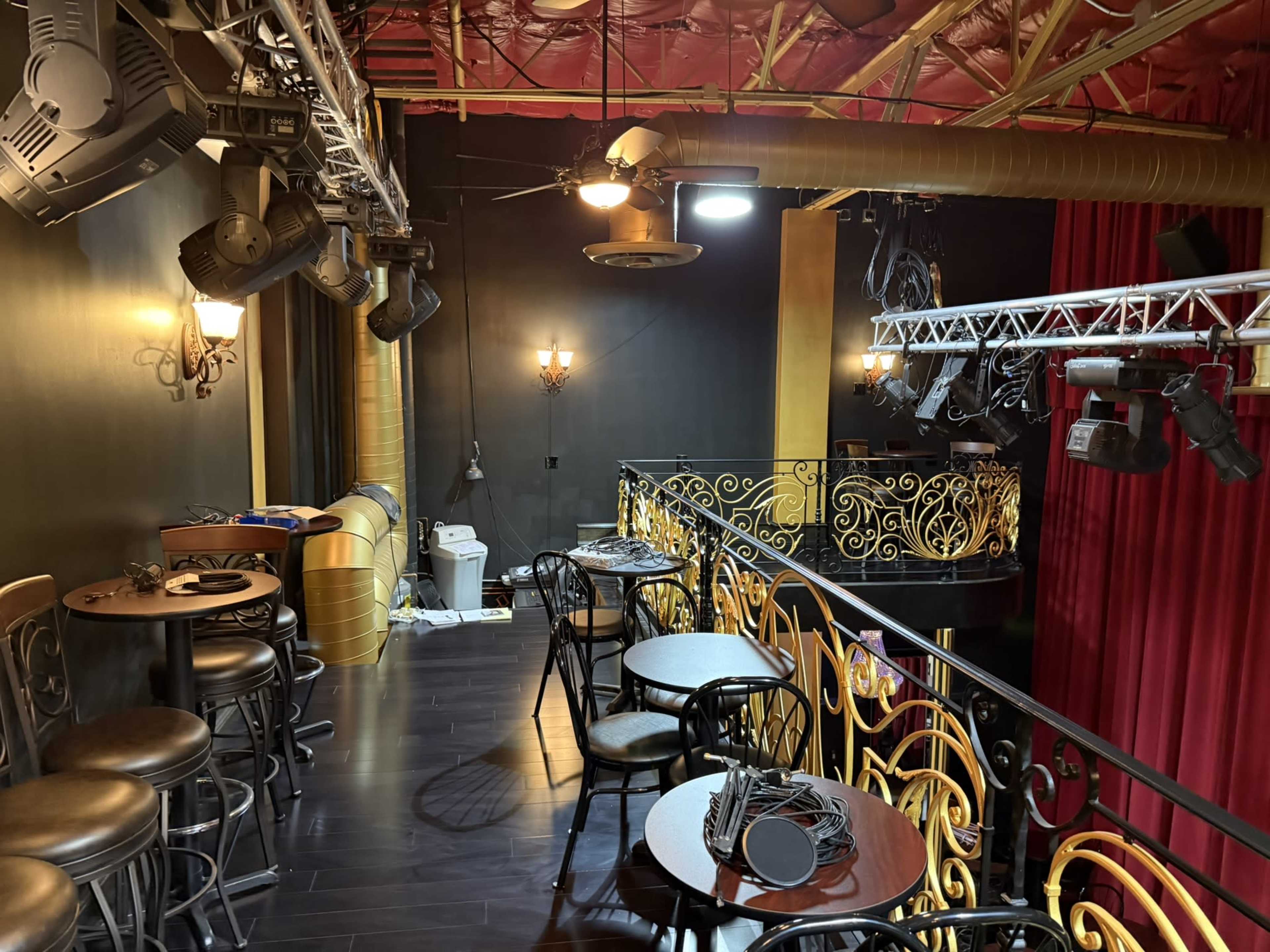The image shows a small lounge area with several round tables and chairs, overlooking a performance space adorned with red curtains and stage lighting.