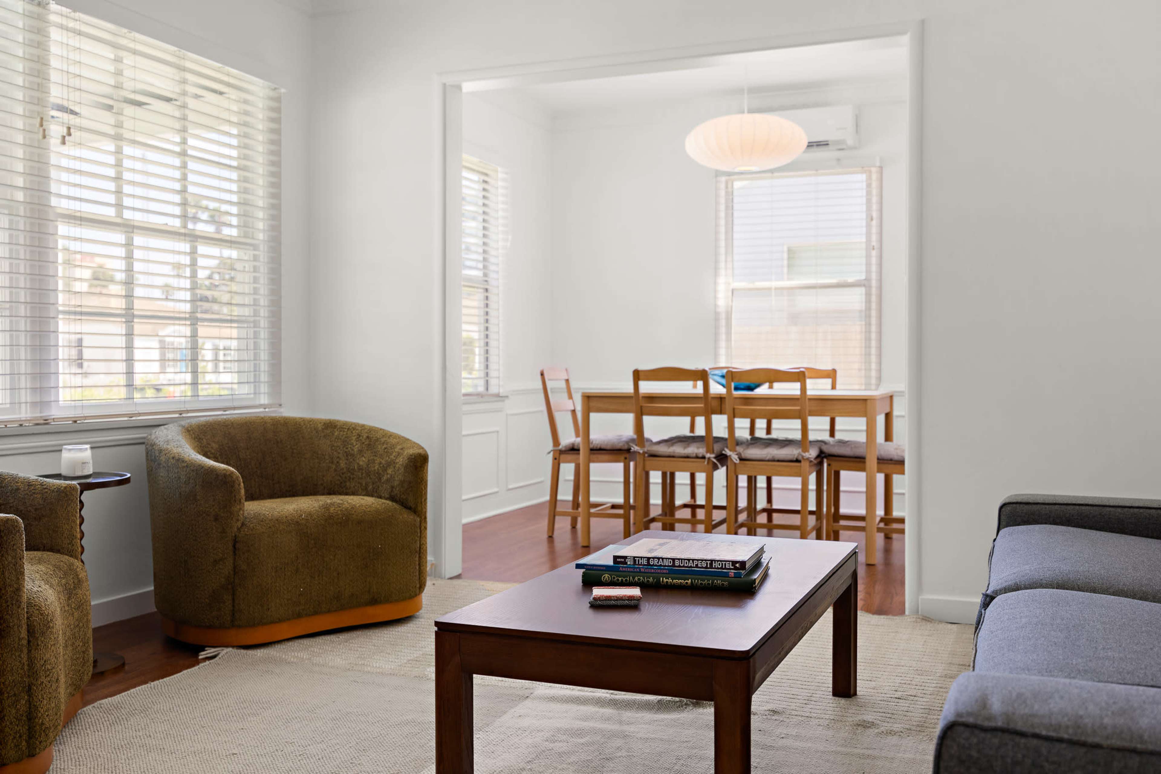 The image shows a living room with two armchairs, a coffee table, and a view into a dining area with a wooden table and chairs framed by white walls and bright windows.