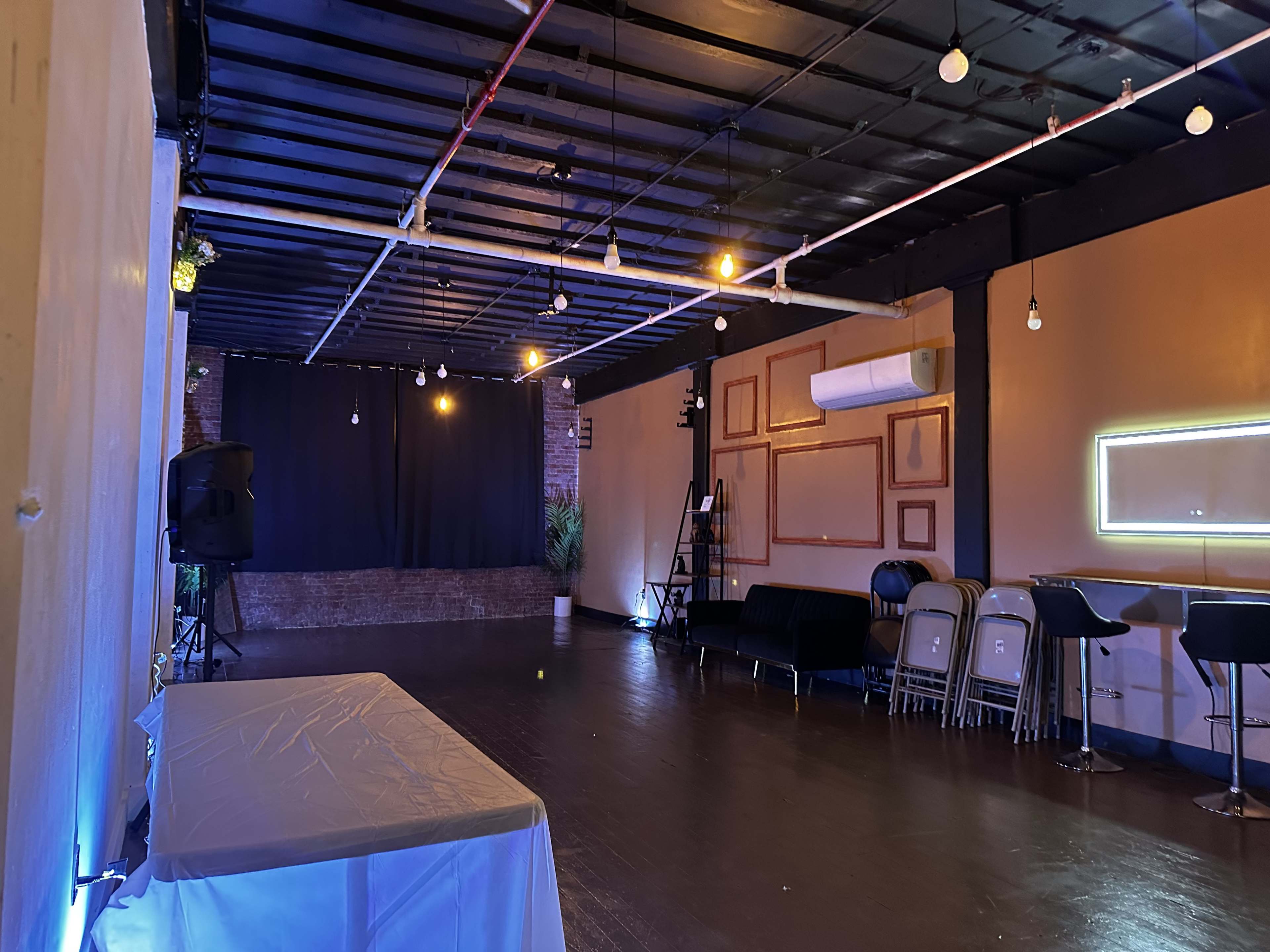 The interior of a dimly lit event space features a black stage area, a white table in the foreground, and rows of metal chairs along the sides.