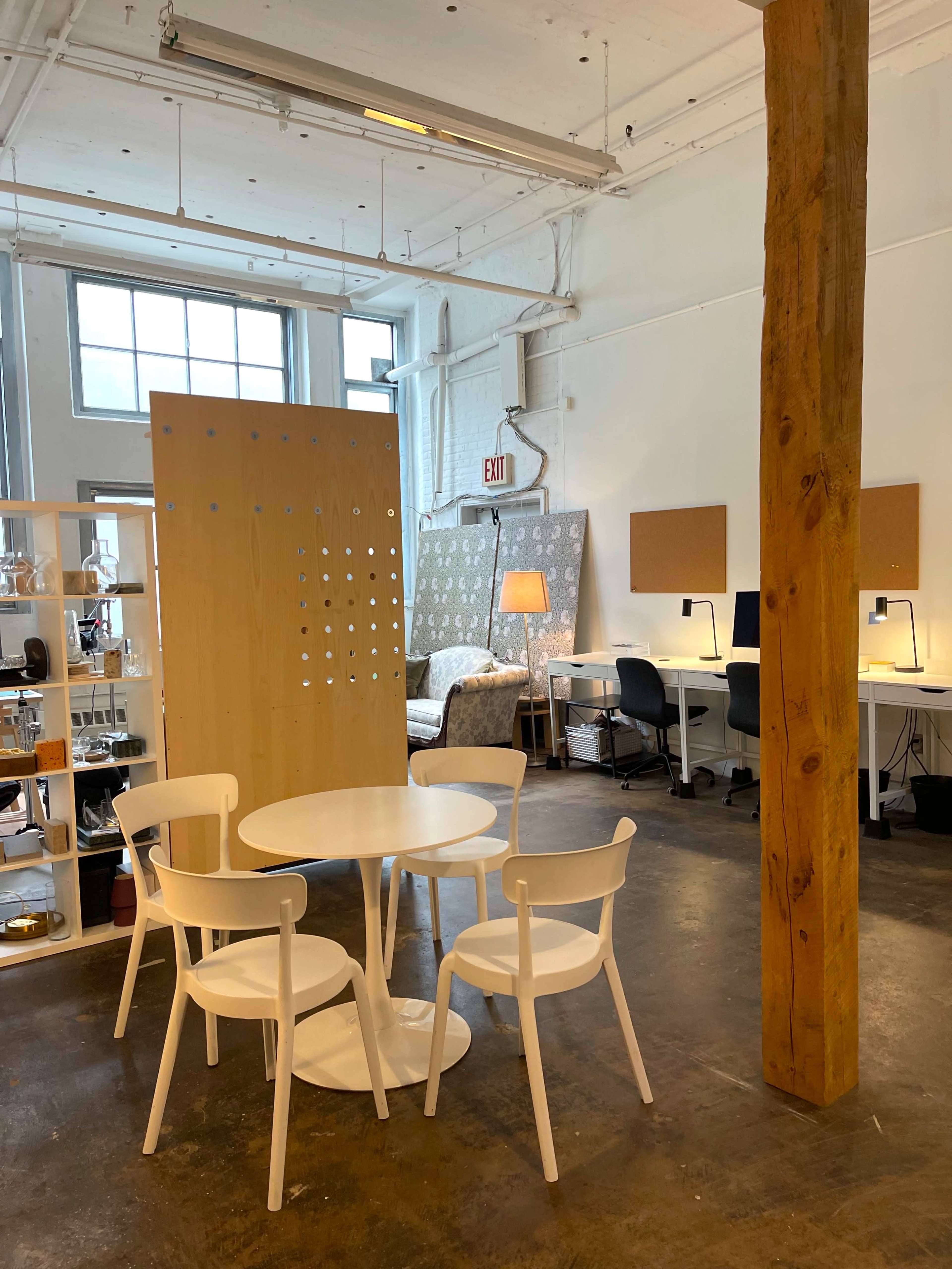 A modern open workspace featuring a small round table with four white chairs, a wooden partition, and a desk area with two chairs and computer monitors in the background.
