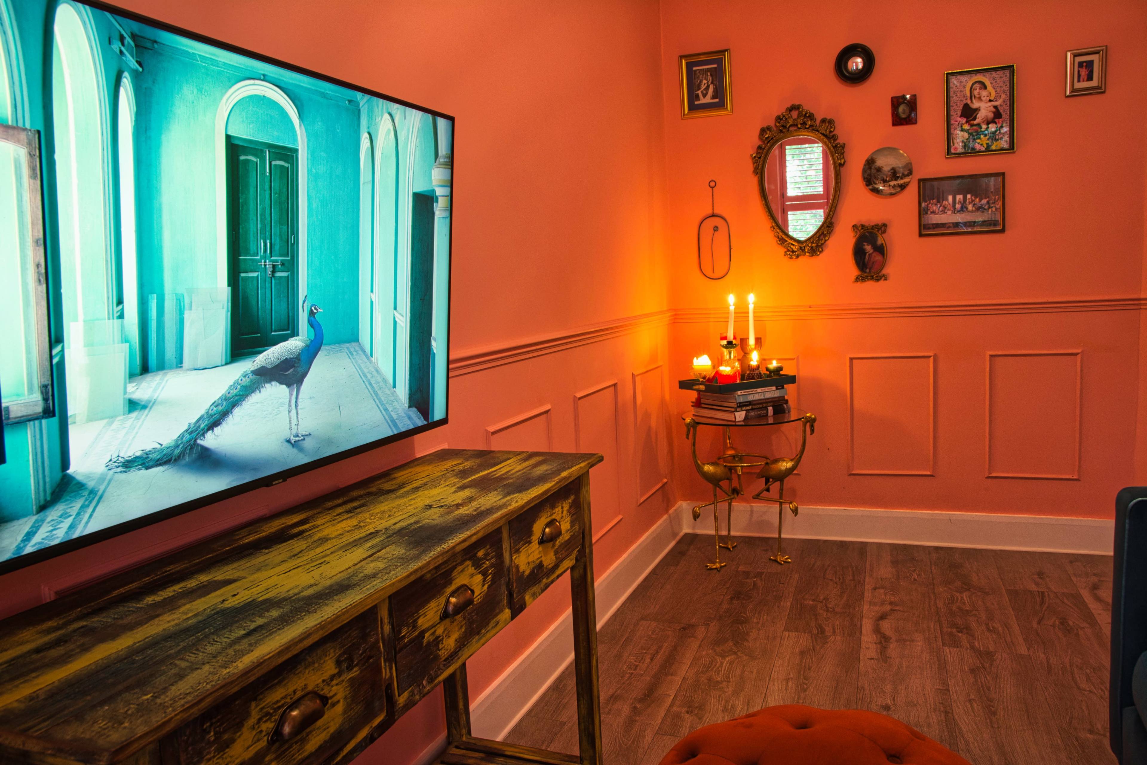 The room features a vintage wooden table, a large television displaying a peacock, and various framed artworks on coral-colored walls, with candles on a small table casting warm light.