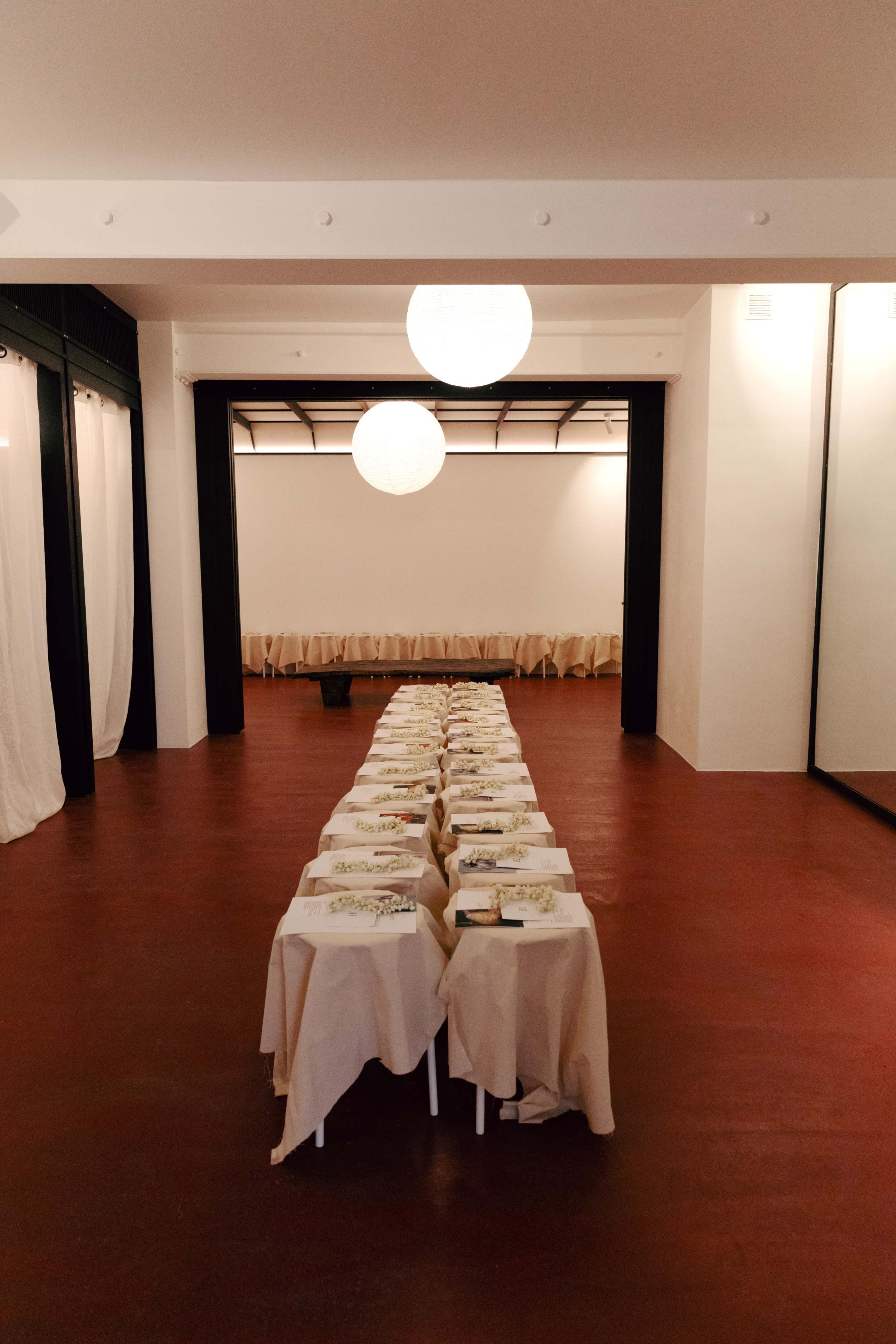 The image shows a spacious dining area with several long tables set up, each covered with white tablecloths and arranged in a line, while large round lanterns hang from the ceiling.