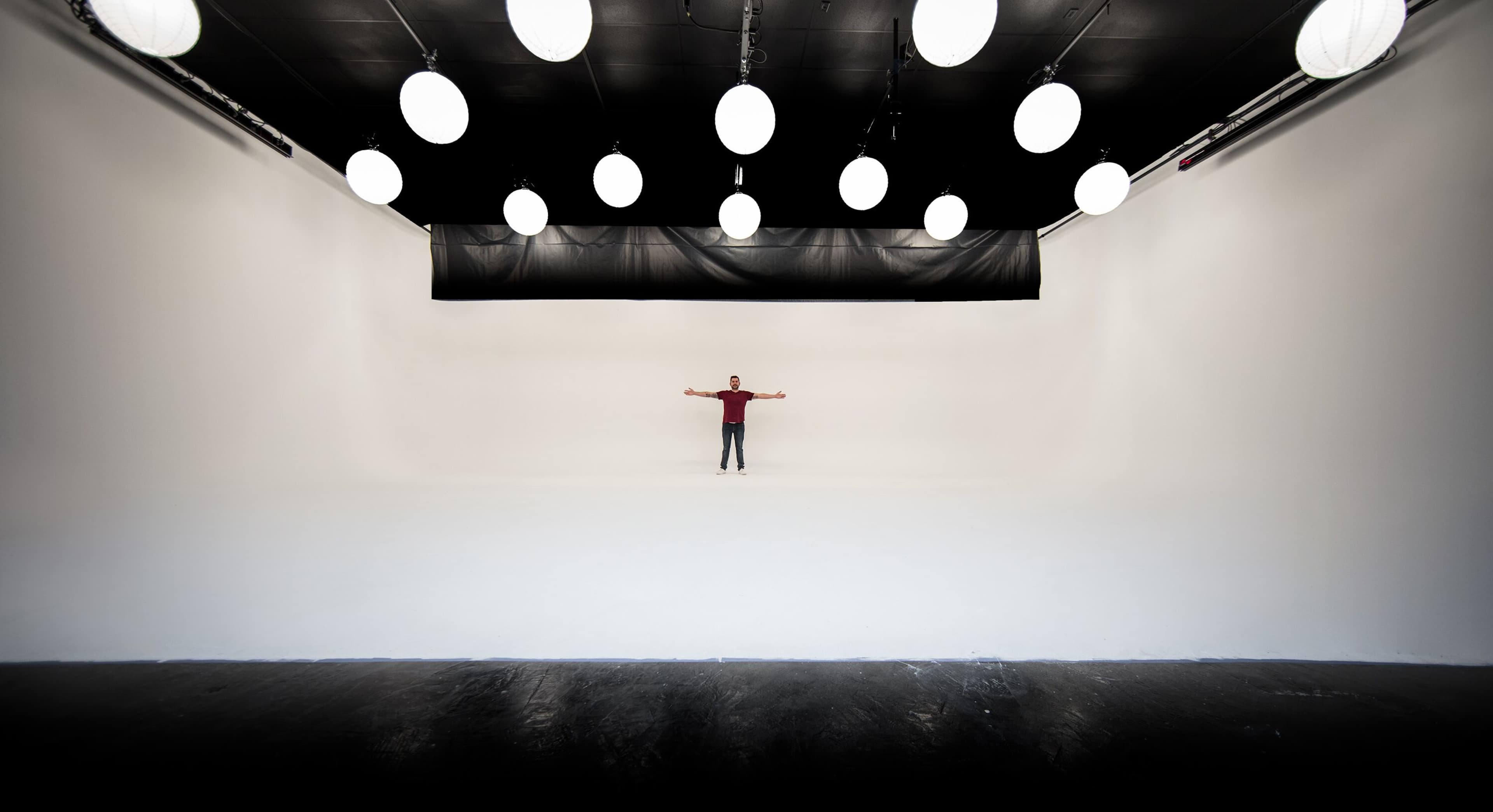 A person stands with arms outstretched in a large, empty studio with white walls and round hanging lights.