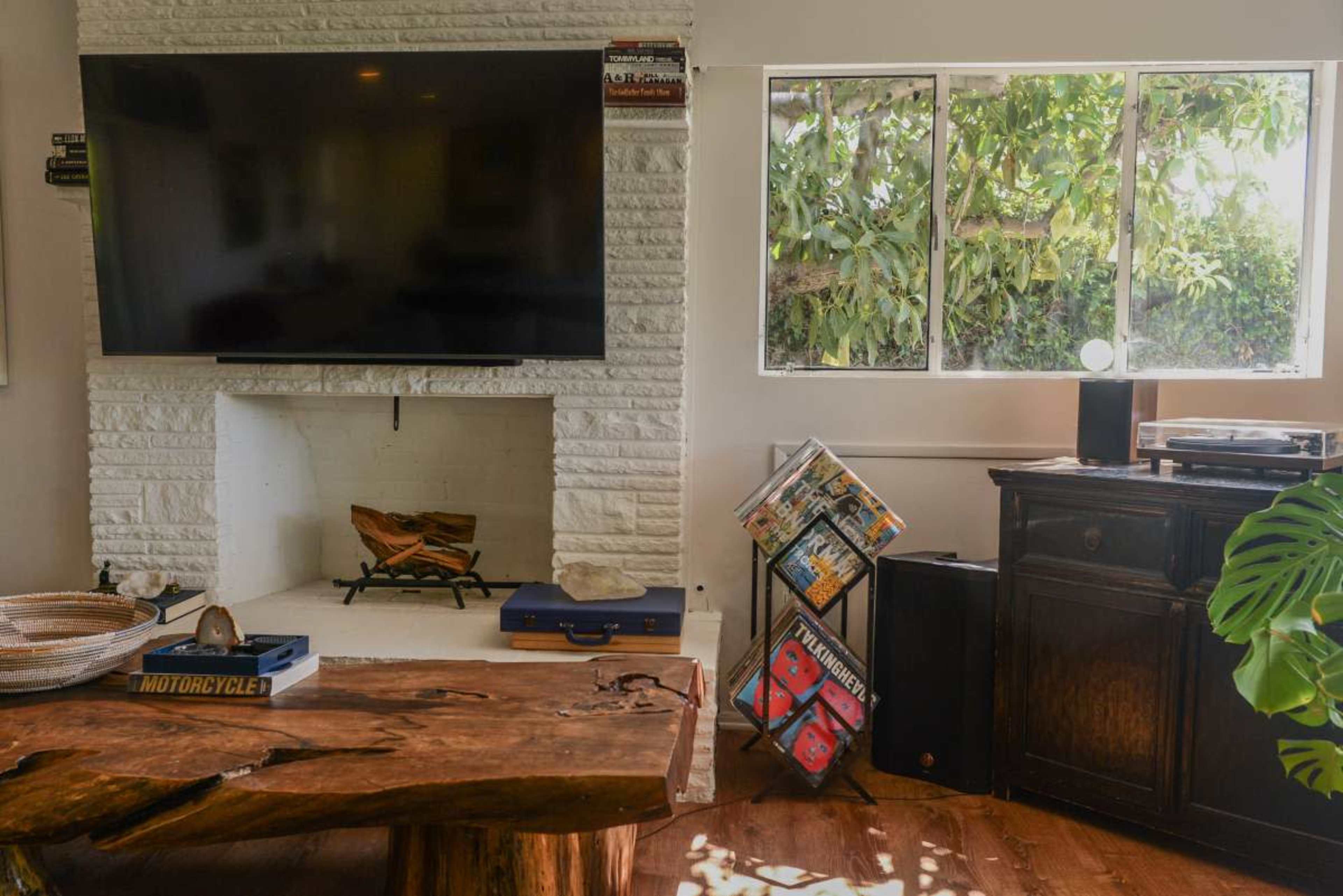The image depicts a living room with a white brick fireplace, a large television mounted above it, and a wooden table in the foreground, alongside a record player and a collection of magazines on display.
