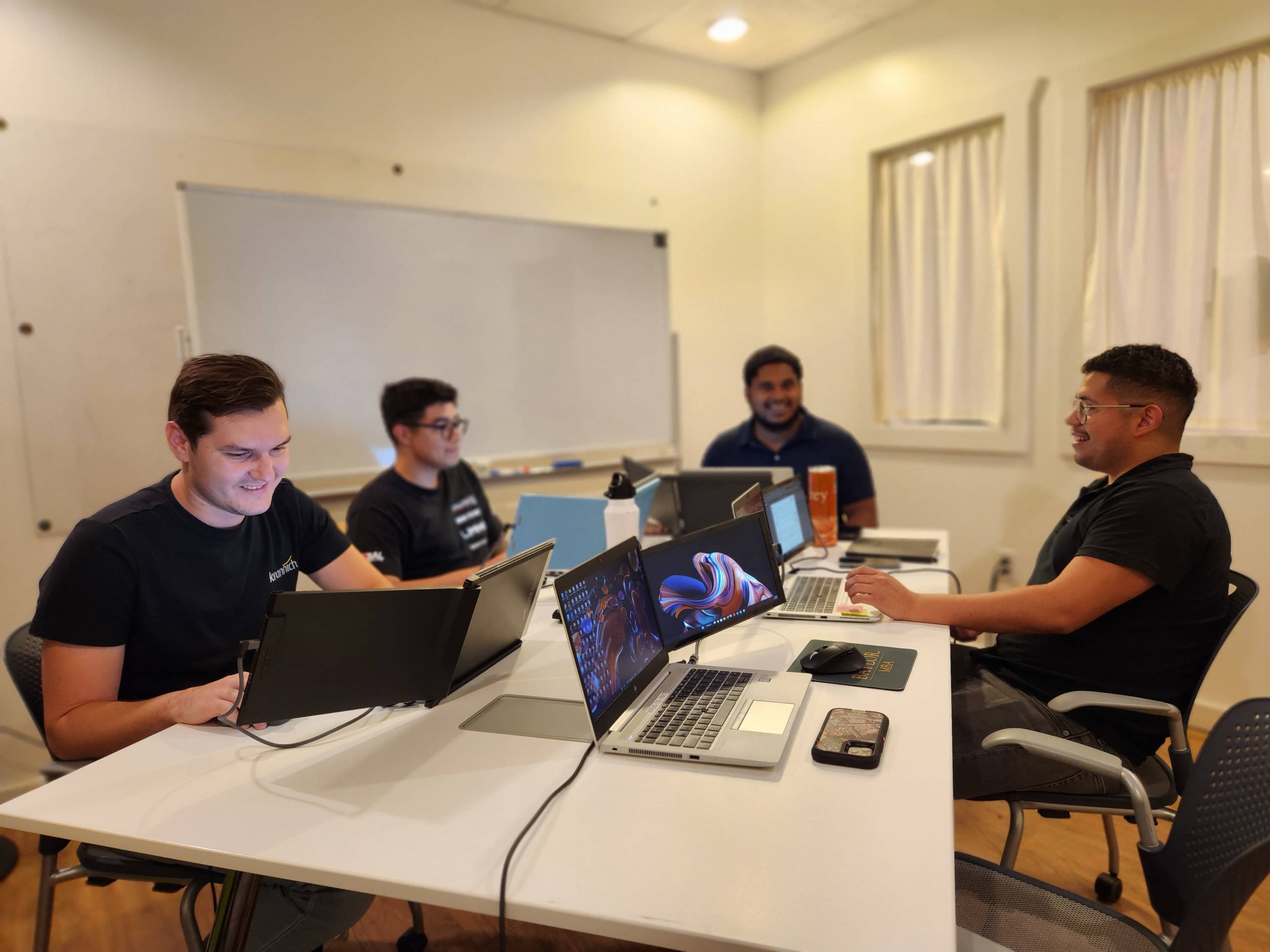 Four individuals are seated around a table in a well-lit room, each using laptops and engaged in a collaborative work session.
