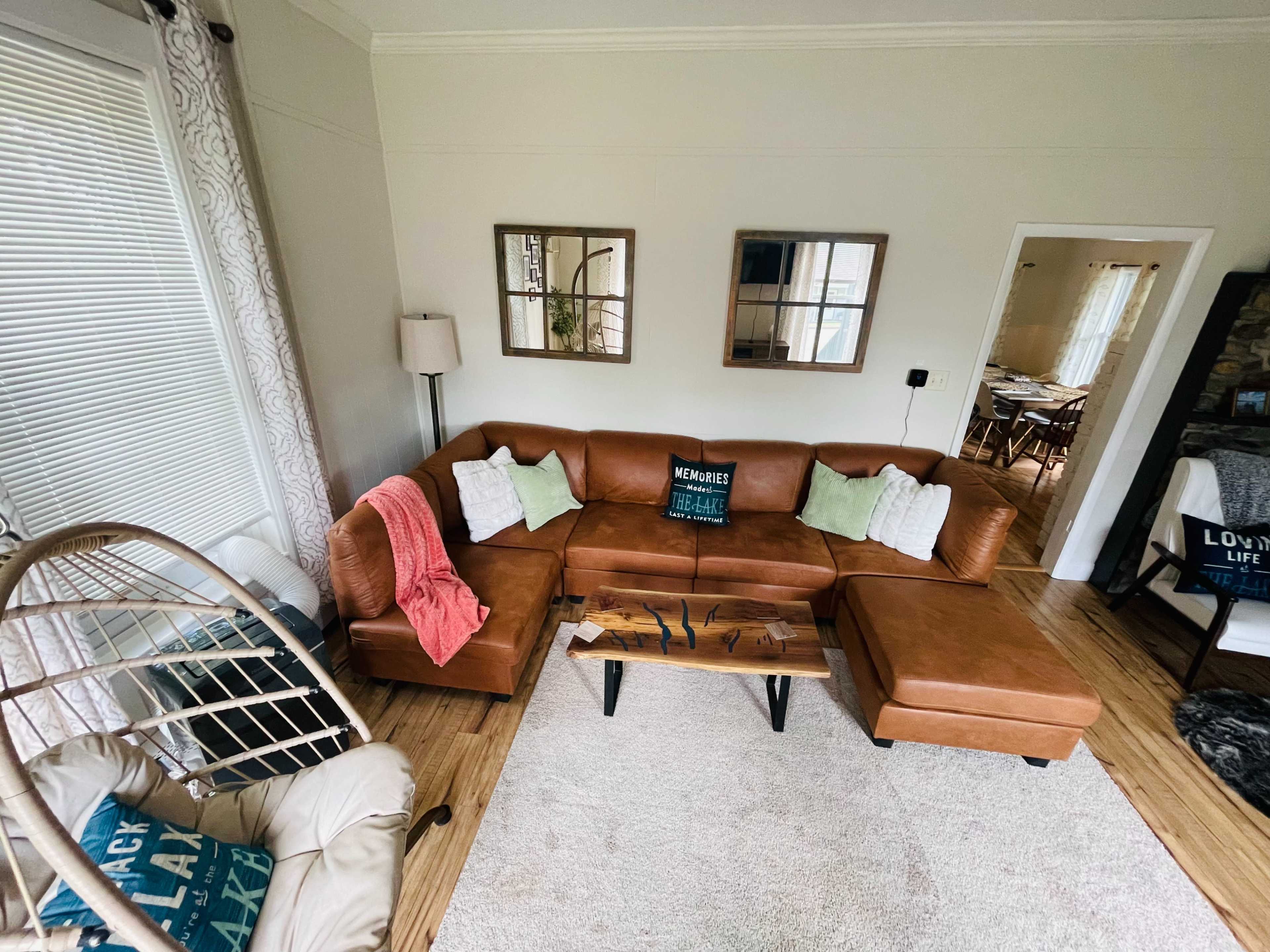A cozy living room with a brown leather sectional couch, a wooden coffee table, and two mirrors on the wall, surrounded by light-colored walls and a soft rug.