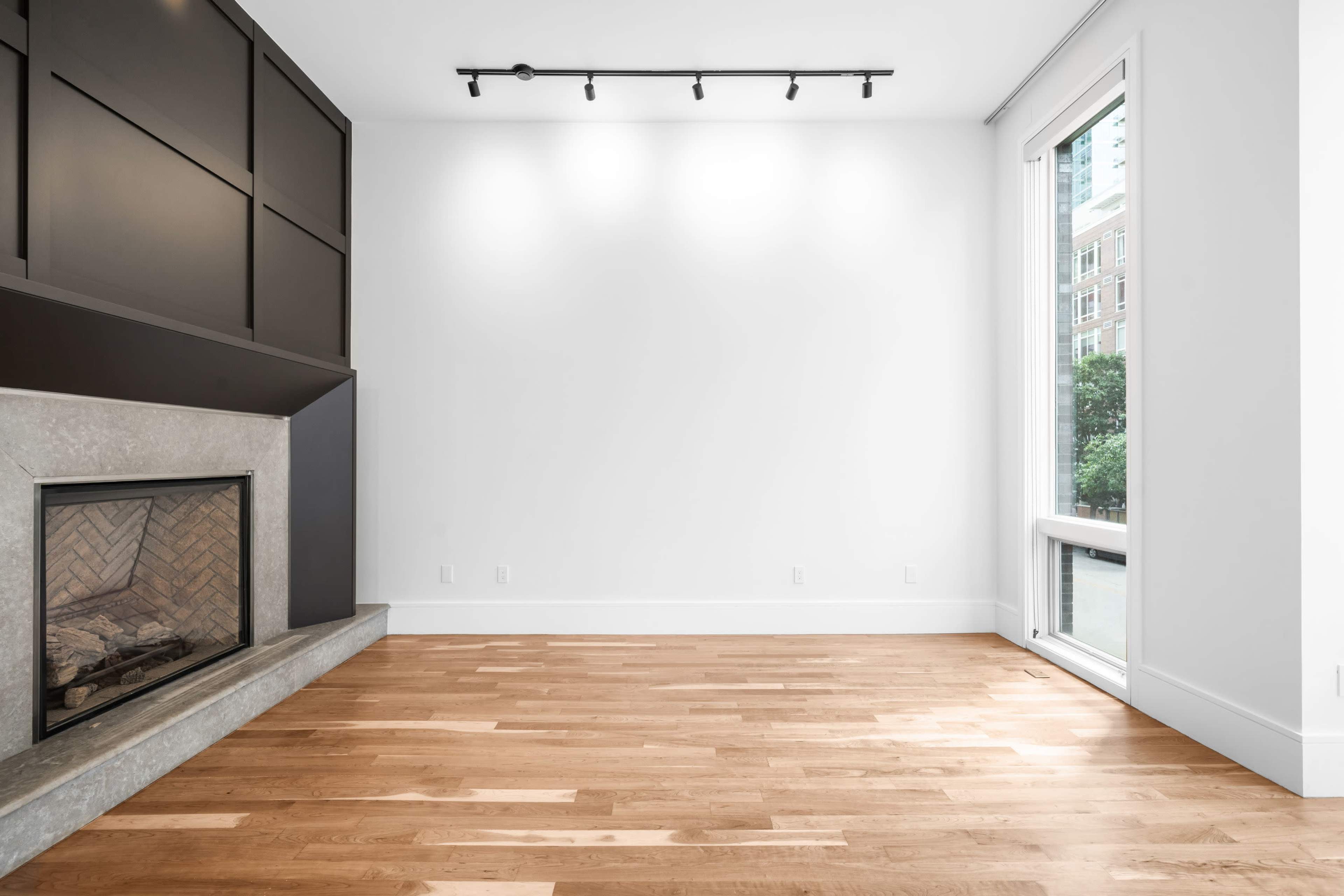 The image shows a spacious, empty room featuring a modern fireplace and a large window with natural light entering.