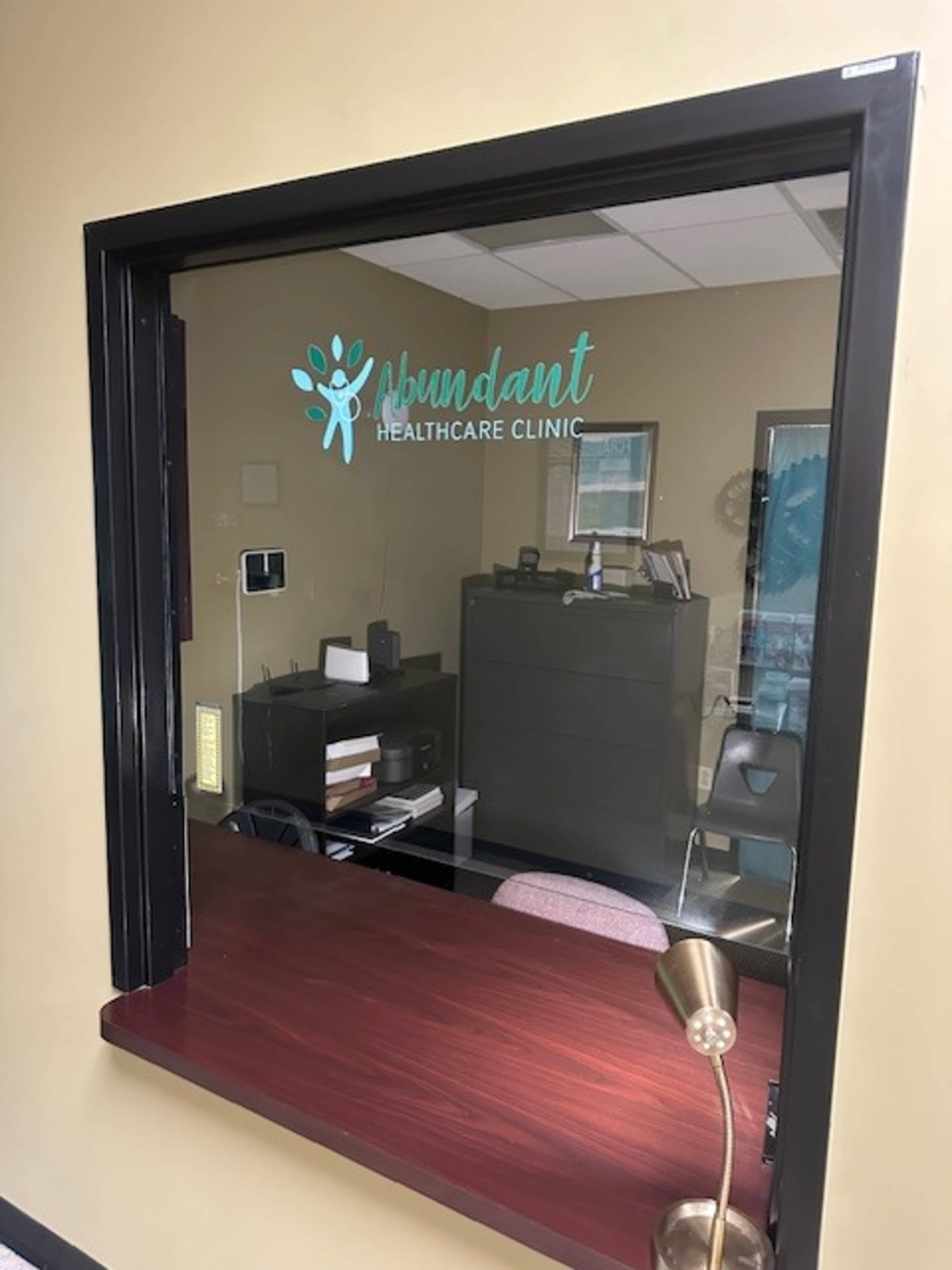 The image shows a glass window with the words "Abundant Healthcare Clinic" displayed above a reception area with a desk and office furniture visible behind it.