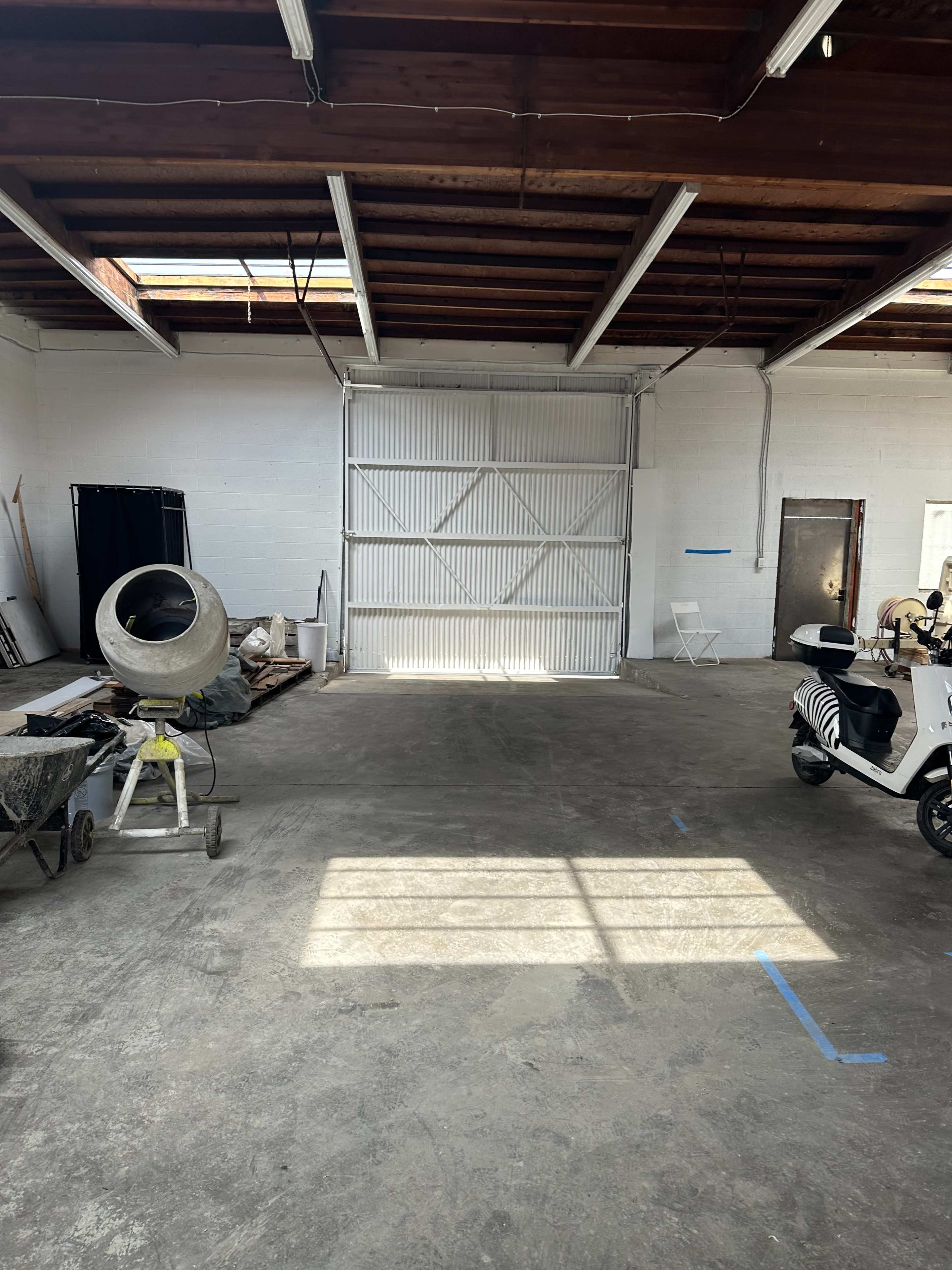 The image shows an empty warehouse interior with concrete flooring, a large metal door at one end, and construction equipment scattered throughout.