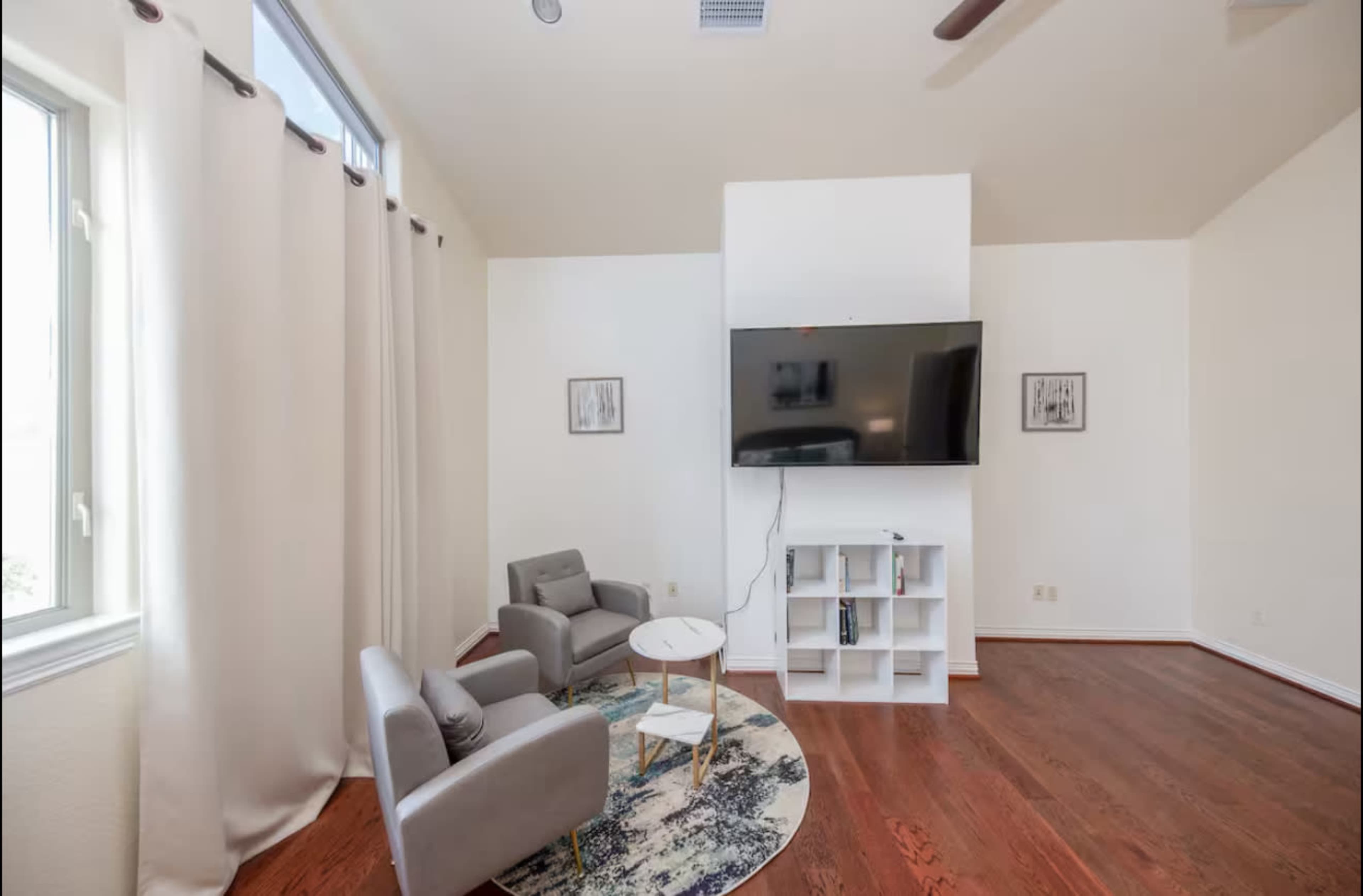 The image shows a minimalist living room with two gray armchairs, a small white table, a television mounted on a white wall, and a circular rug.