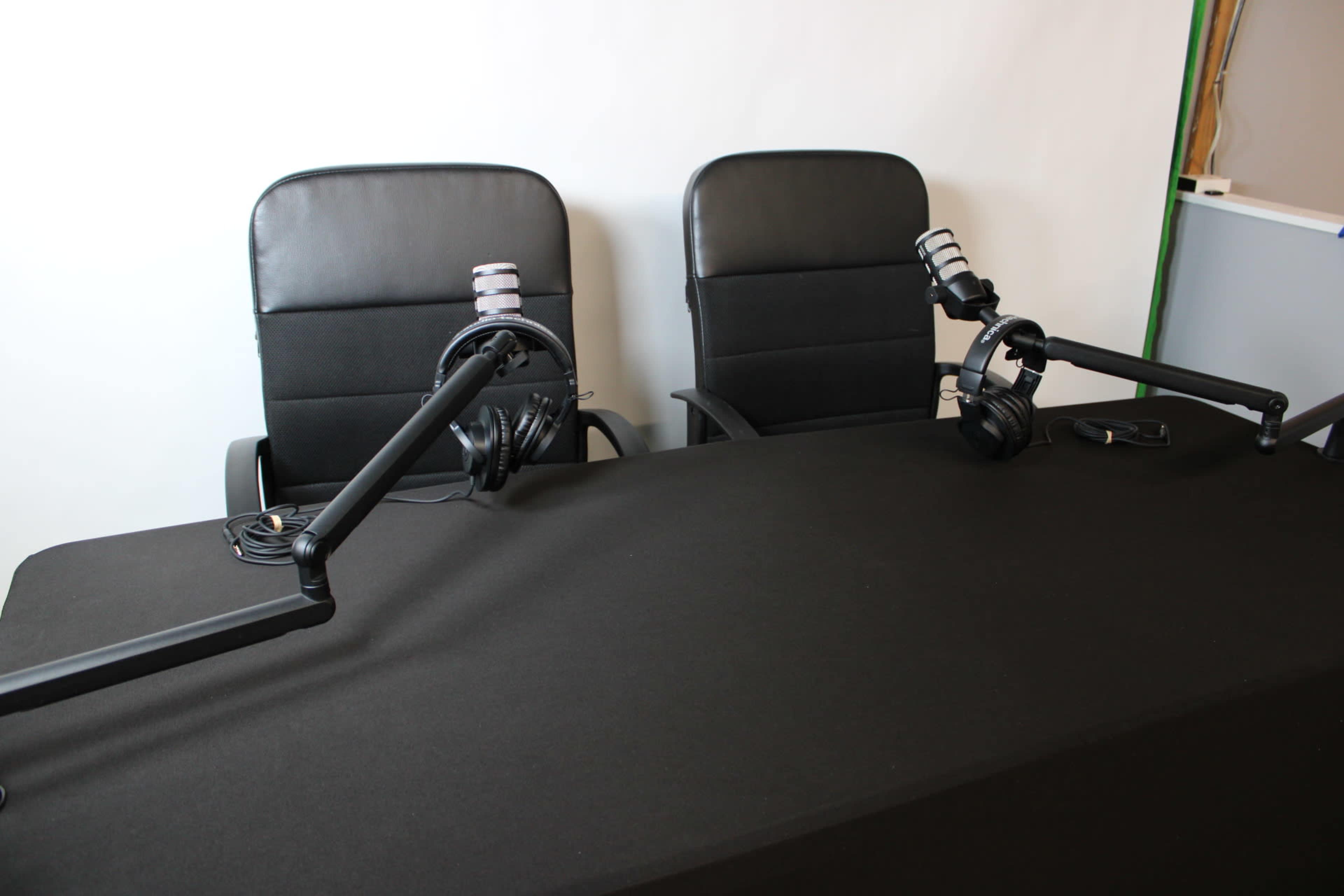 The image shows two black office chairs positioned at a table draped with a black cloth, each equipped with a microphone and headphones.
