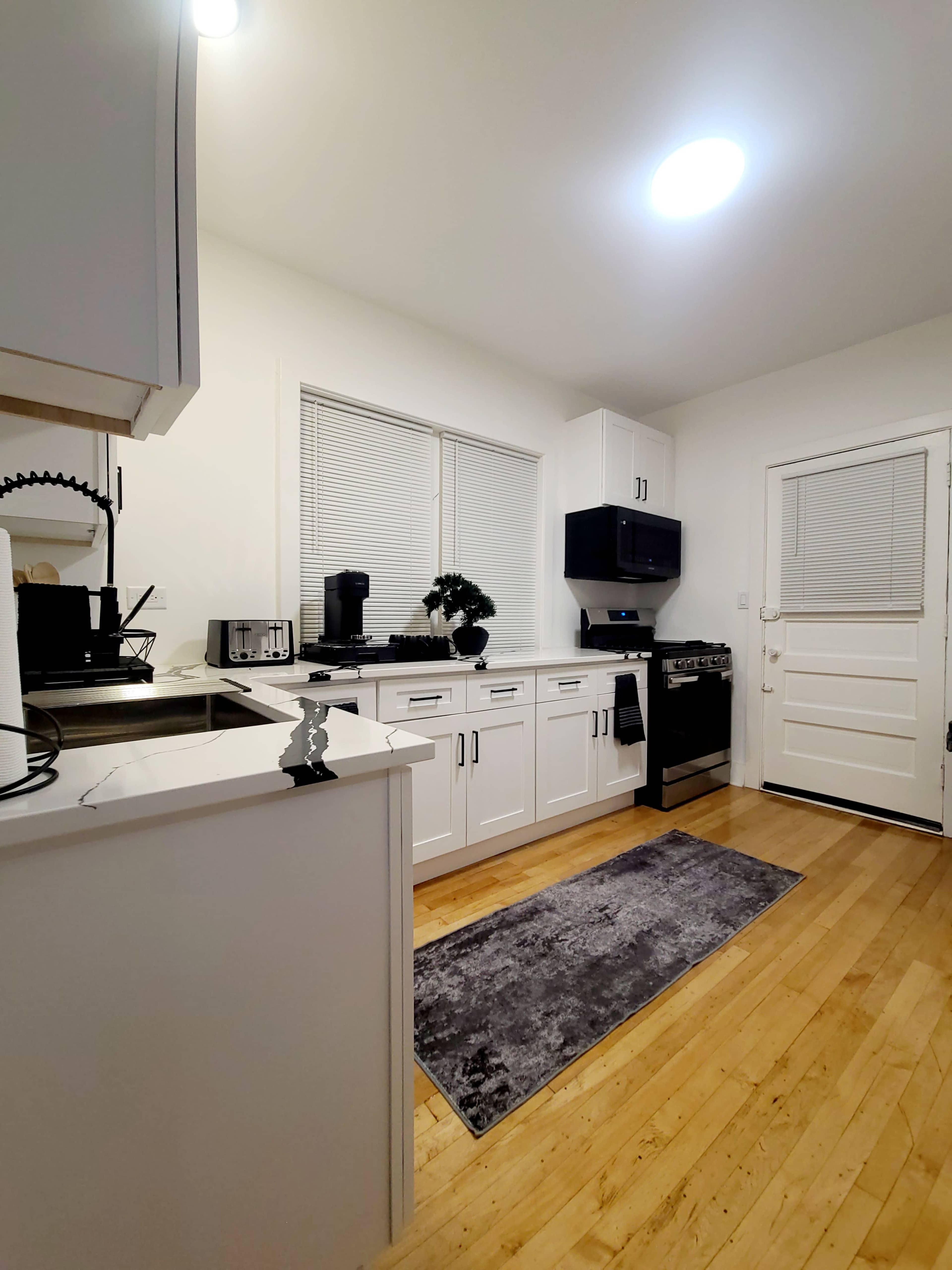 The image shows a modern kitchen with white cabinets, stainless steel appliances, a gray rug, and a door leading outside.