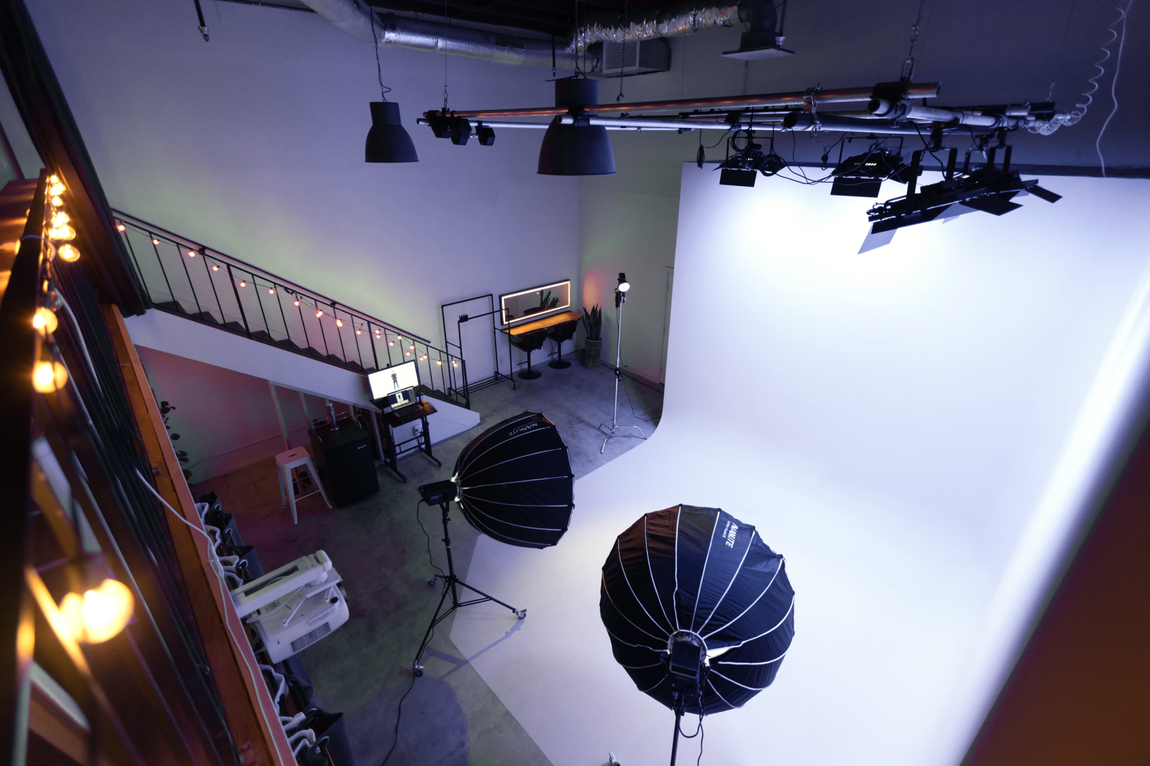 A photography studio featuring a large white backdrop, two softbox lights, and a staircase with string lights in the background.